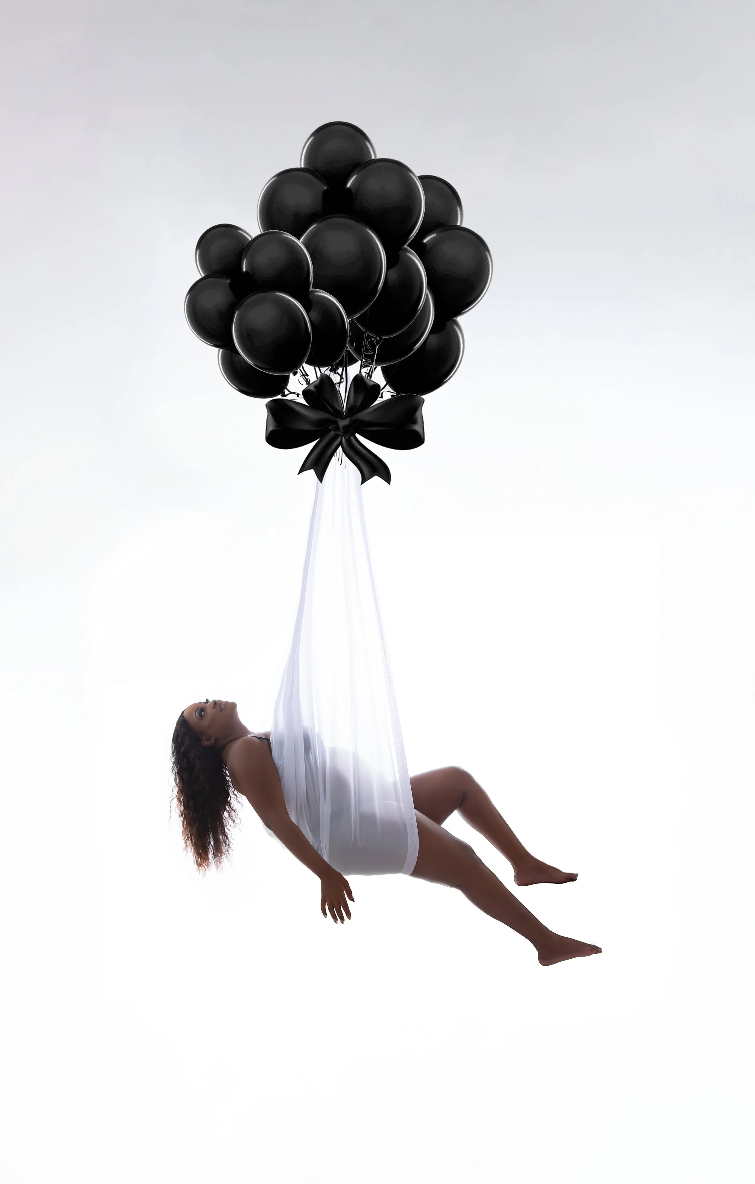 A woman in a white dress lying backwards while suspended by black balloons tied with a black ribbon, against a white background.