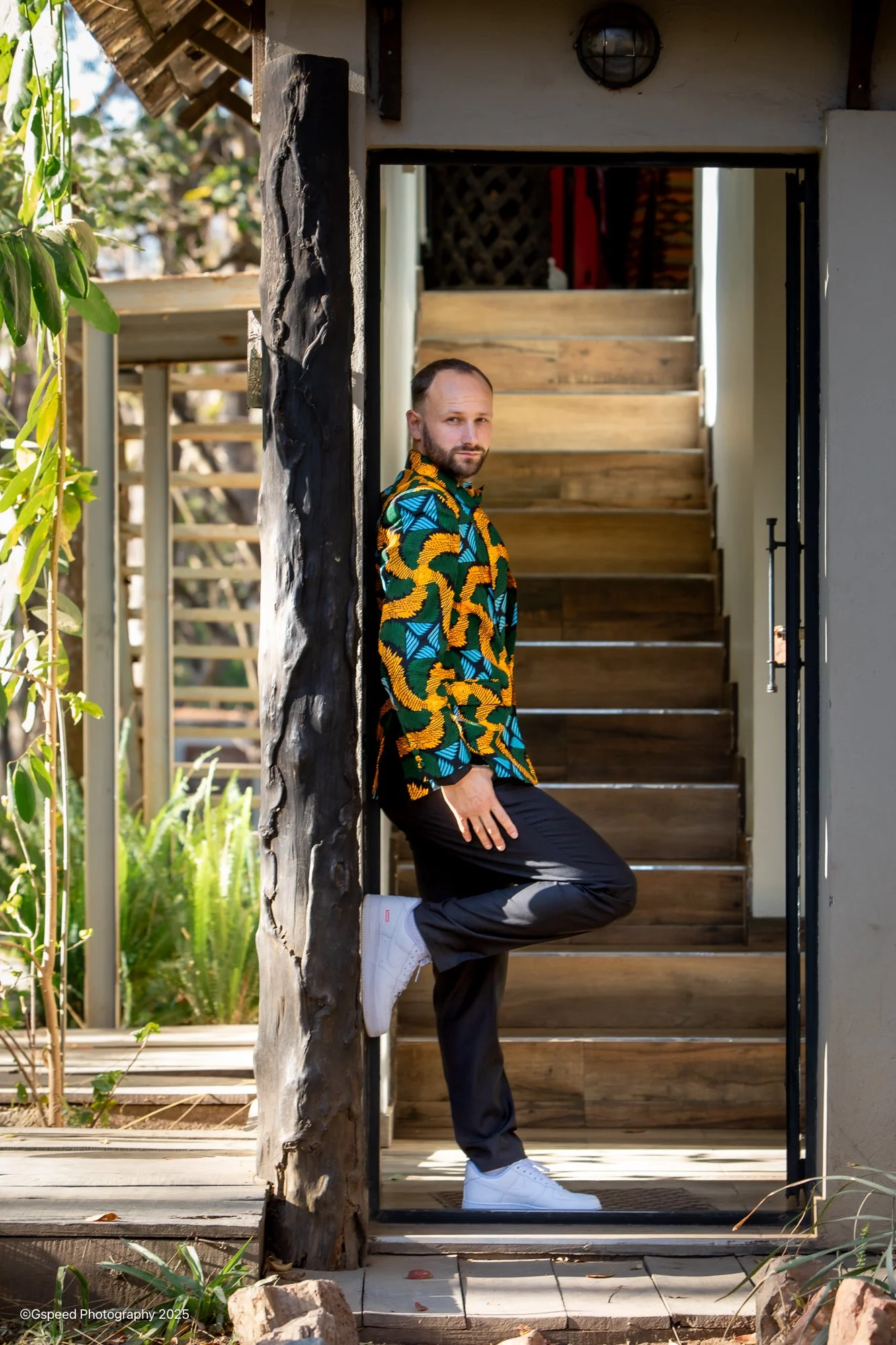 A man in a colorful jacket and black pants leaning against a wooden post outside, standing on one leg with the other bent and foot resting on the ground, near a staircase and surrounded by plants.