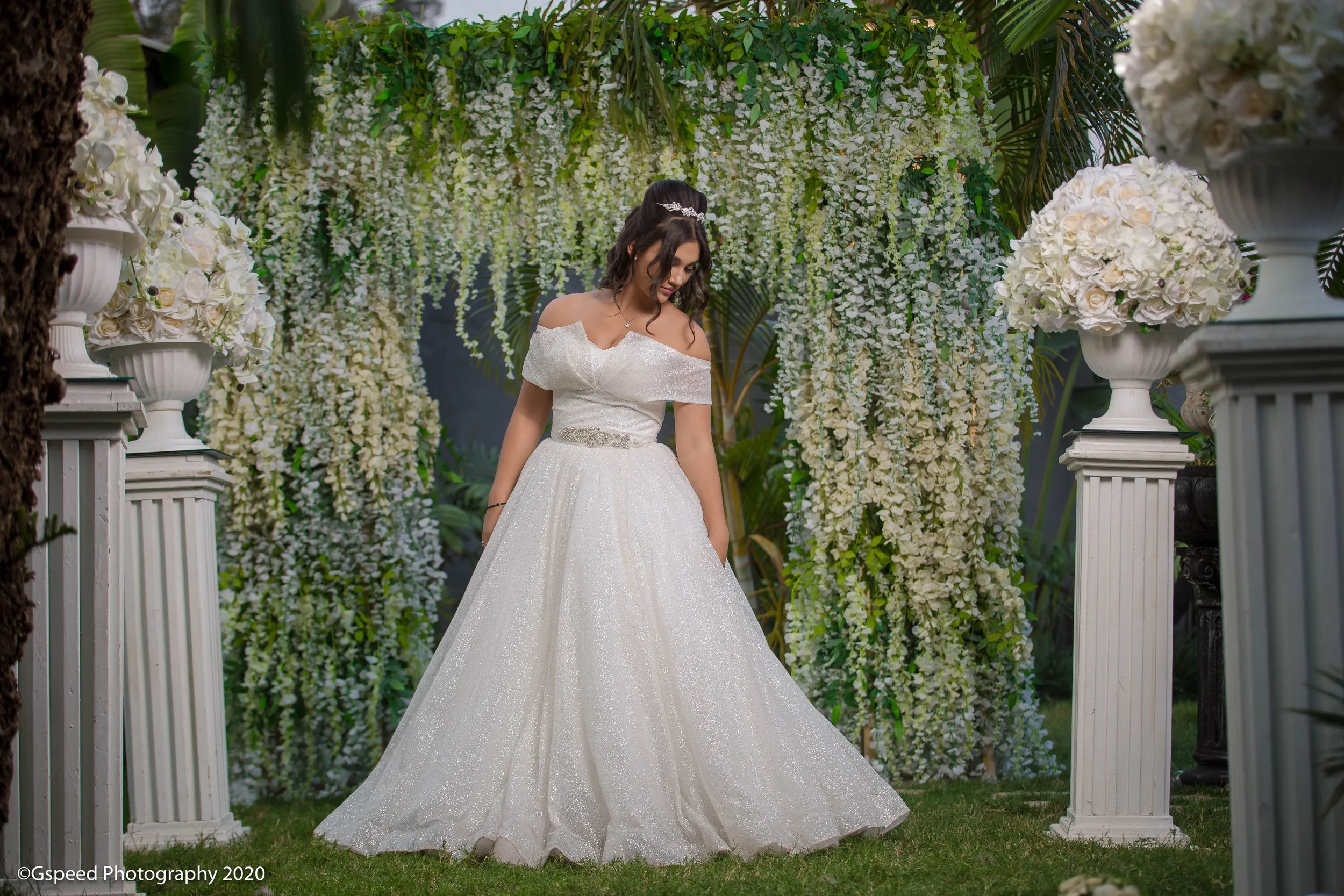 A bride in an off-shoulder white wedding gown with a glittering belt, standing outdoors in front of hanging white flowers and green foliage, surrounded by white floral arrangements on pedestals and white picket fences.