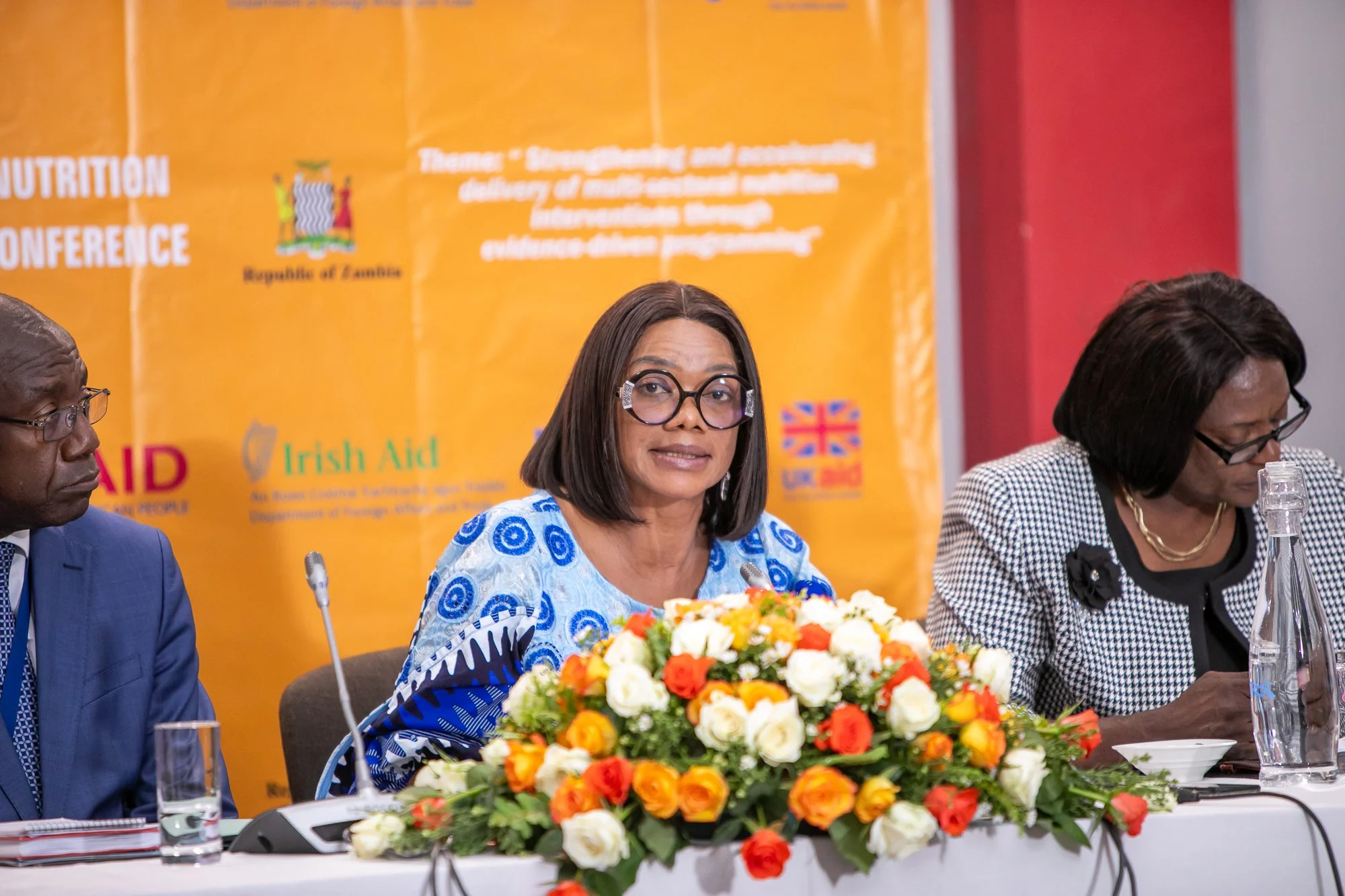 Three people seated at a conference table with a large bouquet of orange, white, and red flowers in front of them, in front of a yellow backdrop with the words 'Nutrition Conference' and the emblem of the Republic of Zambia.