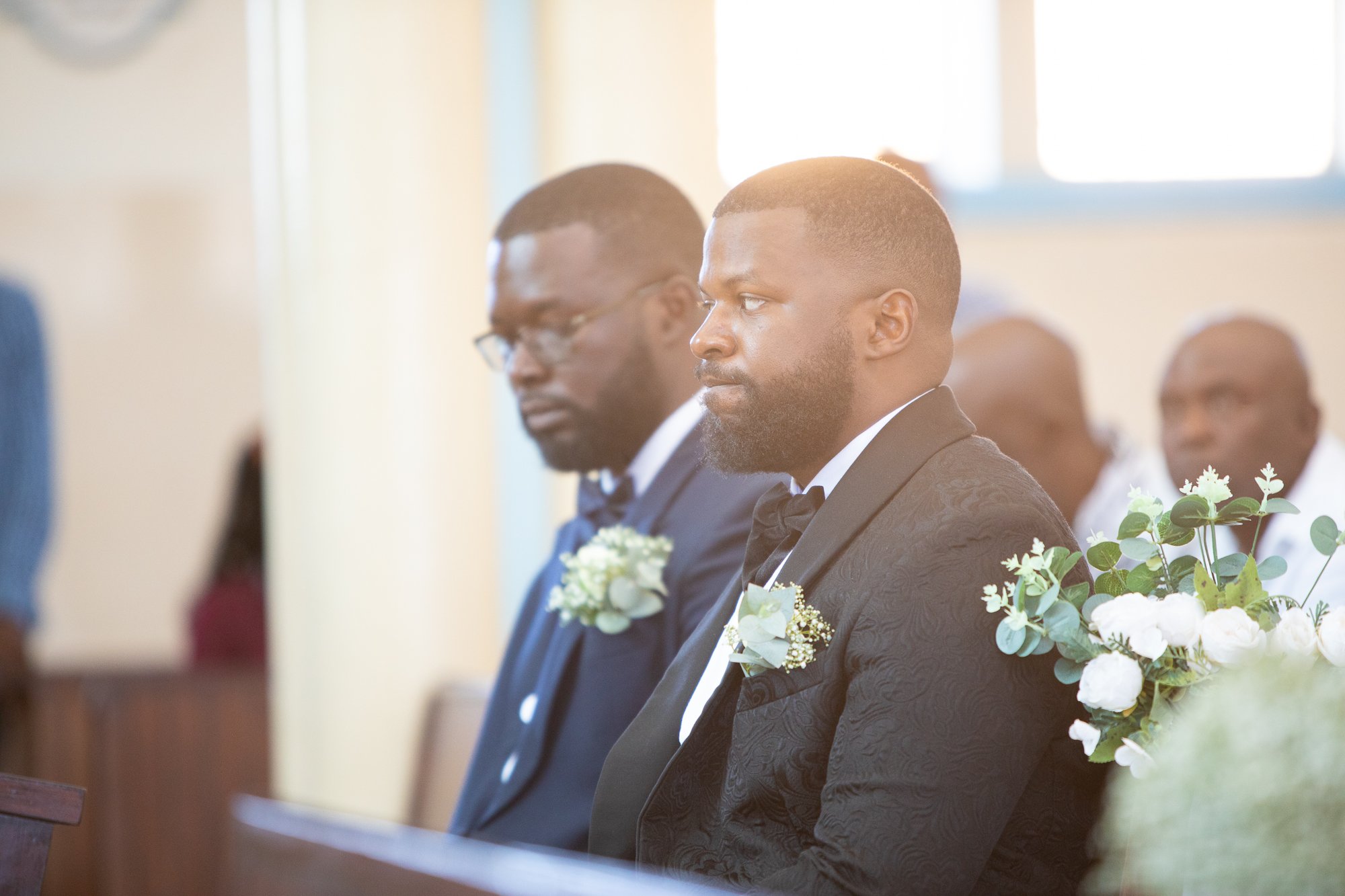 A group of men dressed in tuxedos attending a formal event, seated in a row near floral decorations, with soft natural lighting coming from a window in the background.