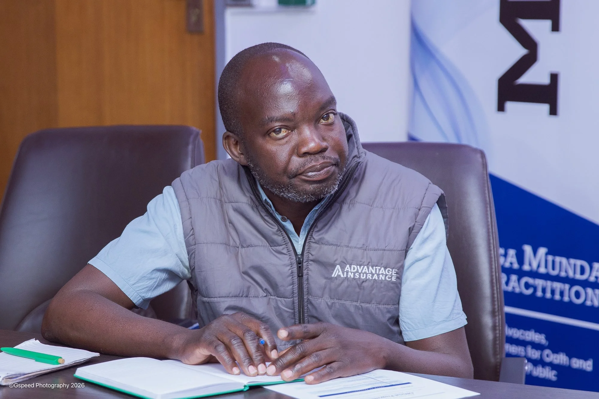 A man with a beard and short hair sitting at a table, wearing a gray vest with the logo "Advantage Insurance." He has a notepad, a green pen, and a blue pen on the table. In the background, there is a partial poster or banner with text that includes 