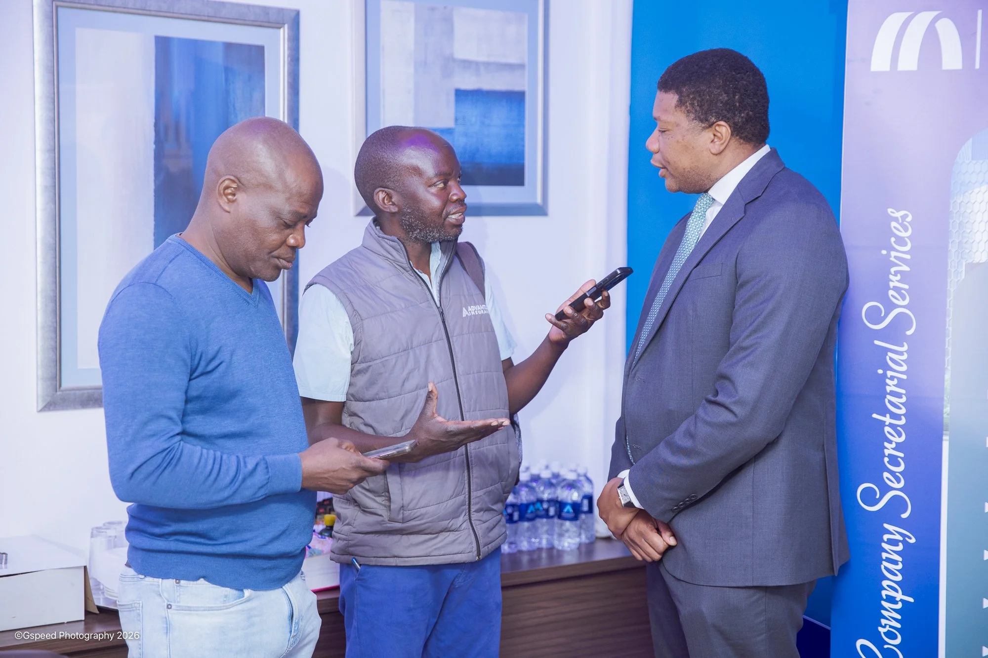 Three men engaged in conversation at an indoor event. One man is in a suit, the other two are dressed casually, holding smartphones. There are water bottles and framed artwork in the background, with a blue banner partially visible on the right.