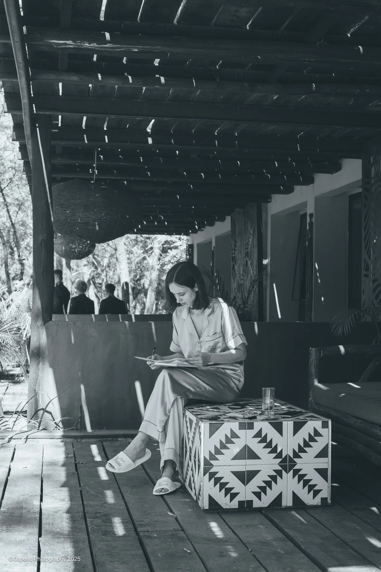 A woman sitting outdoors on a decorative box, reading a book, with sunlight and shadows on the wooden deck and a woman in the background.