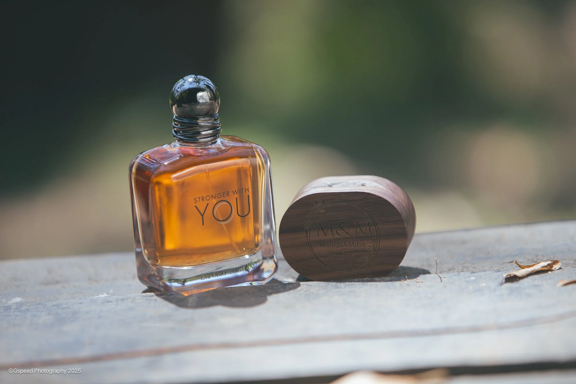 A perfume bottle with an orange liquid labeled 'Stronger With You' and a round wooden container with the initials 'M&M' engraved on the lid, placed on a wooden surface outdoors.