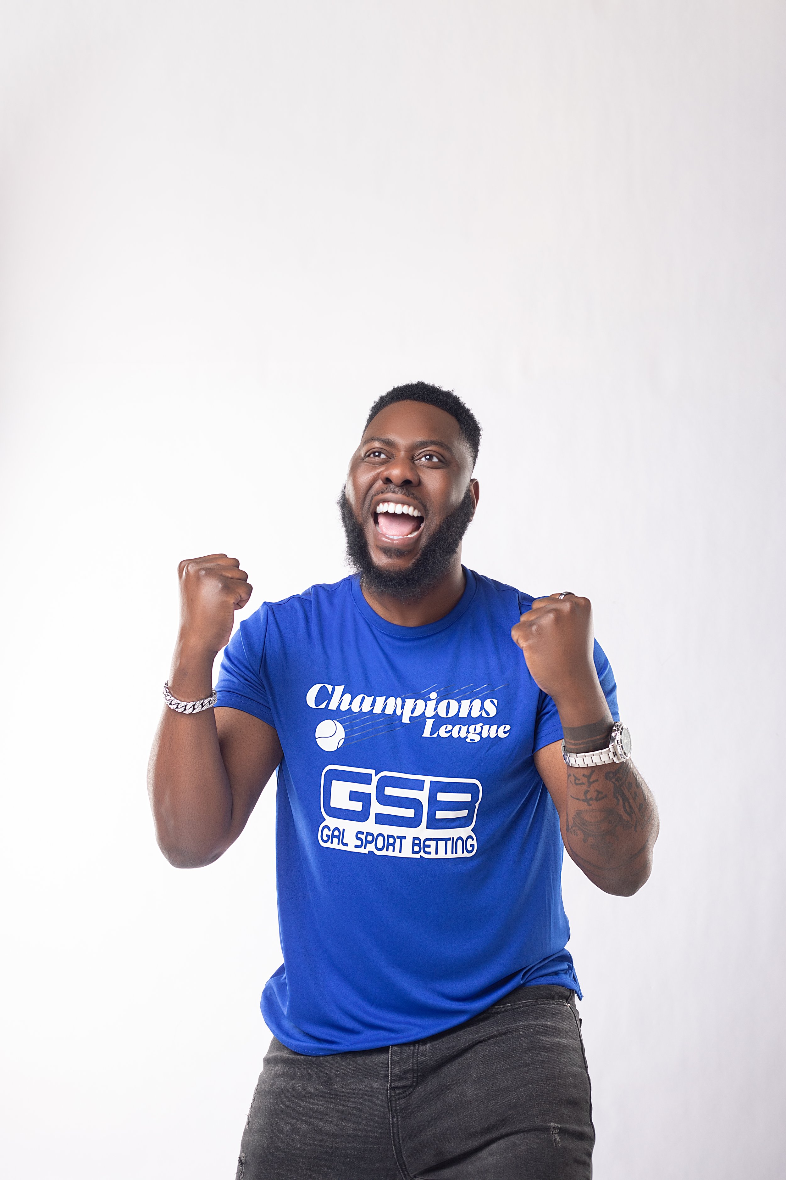 Slap Dee  wearing a blue Champions League T-shirt and gray jeans, showing a victory pose with fists clenched and a wide smile, standing against a plain white background.