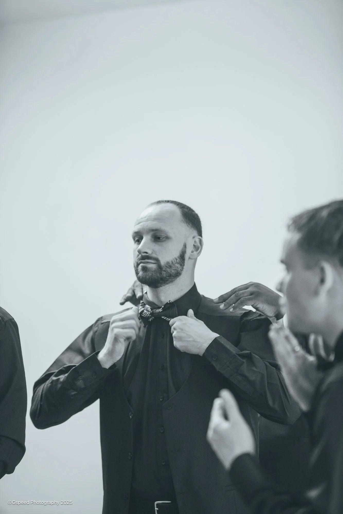 A man in a black dress shirt adjusting a bowtie while others are helping him, in a room with plain white walls.