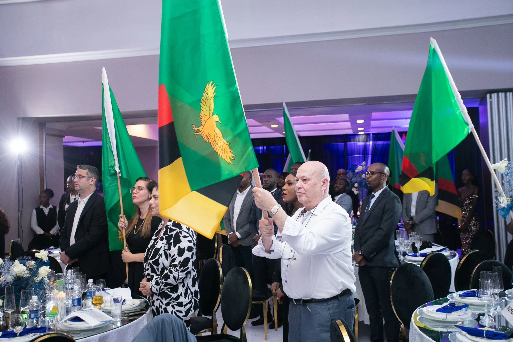 A group of people standing at a banquet table holding flags of Mozambique, which are green, black, yellow, and red with a yellow star and a green emblem. The setting appears to be a formal event or celebration indoors with purple and blue lighting.