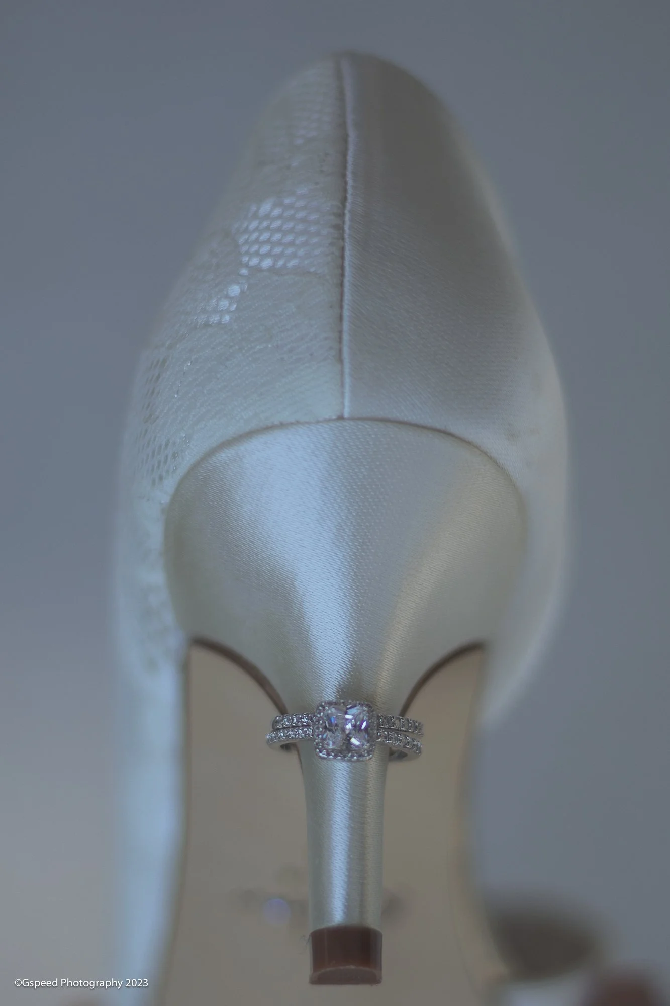 Close-up of a white satin bridal ring pillow with lace and satin detailing. A diamond engagement ring with a halo setting is placed at the front of the pillow. The pillow is set against a plain background.