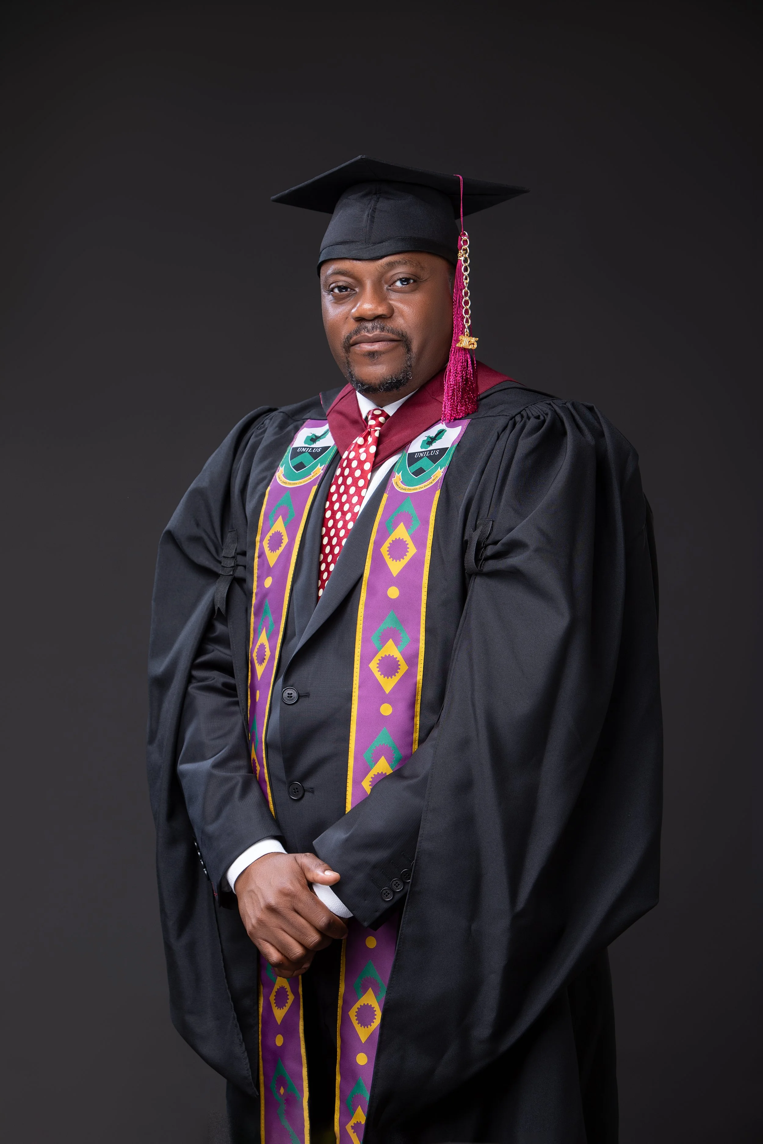A man dressed in academic graduation regalia, including a black cap and gown, with a colorful academic stole, against a dark plain background.