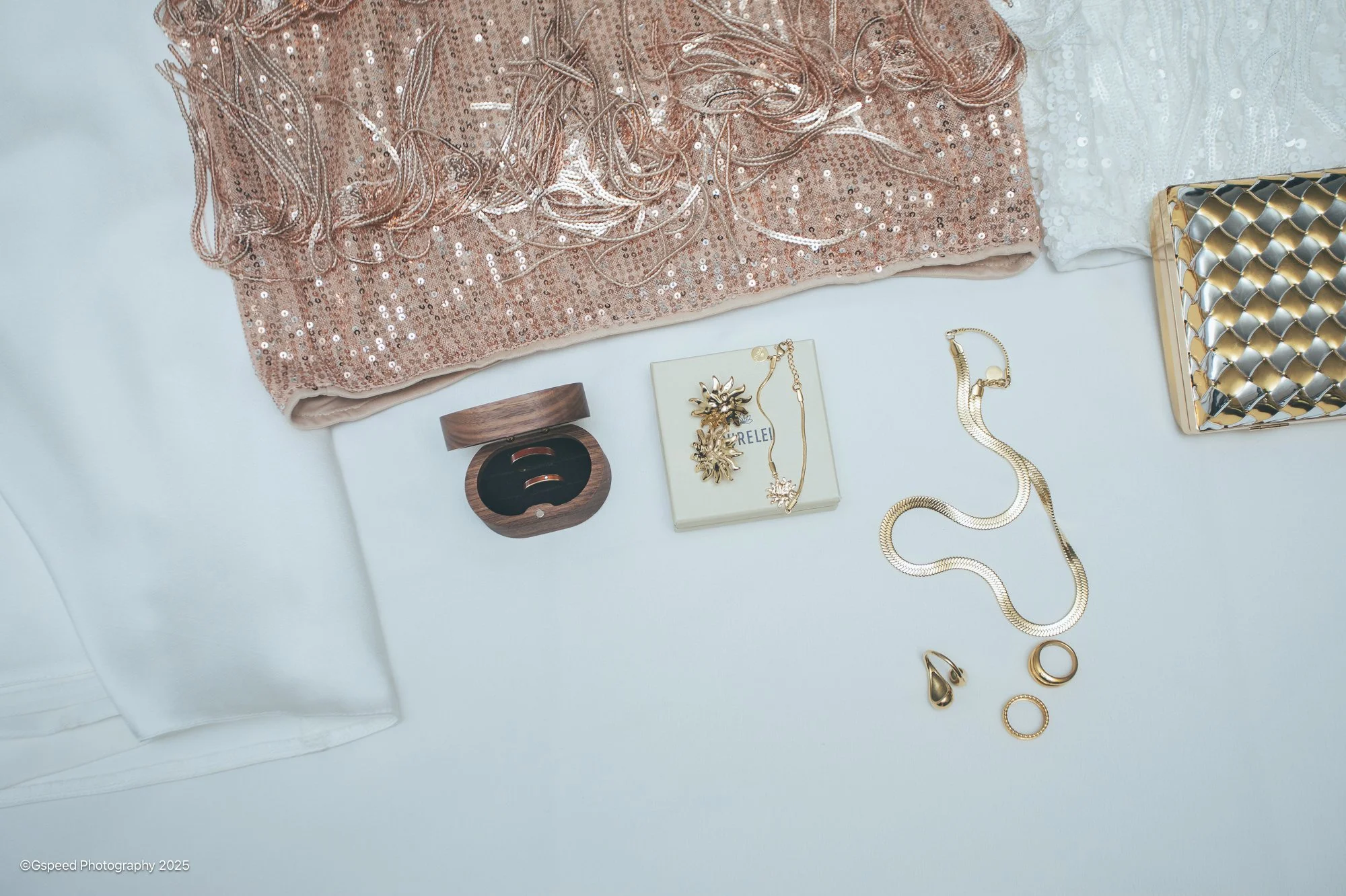 A collection of jewelry and accessories on a white surface, including a pink sequined fabric, a pair of gold earrings, gold and silver necklaces, a gold bracelet, and decorative boxes.
