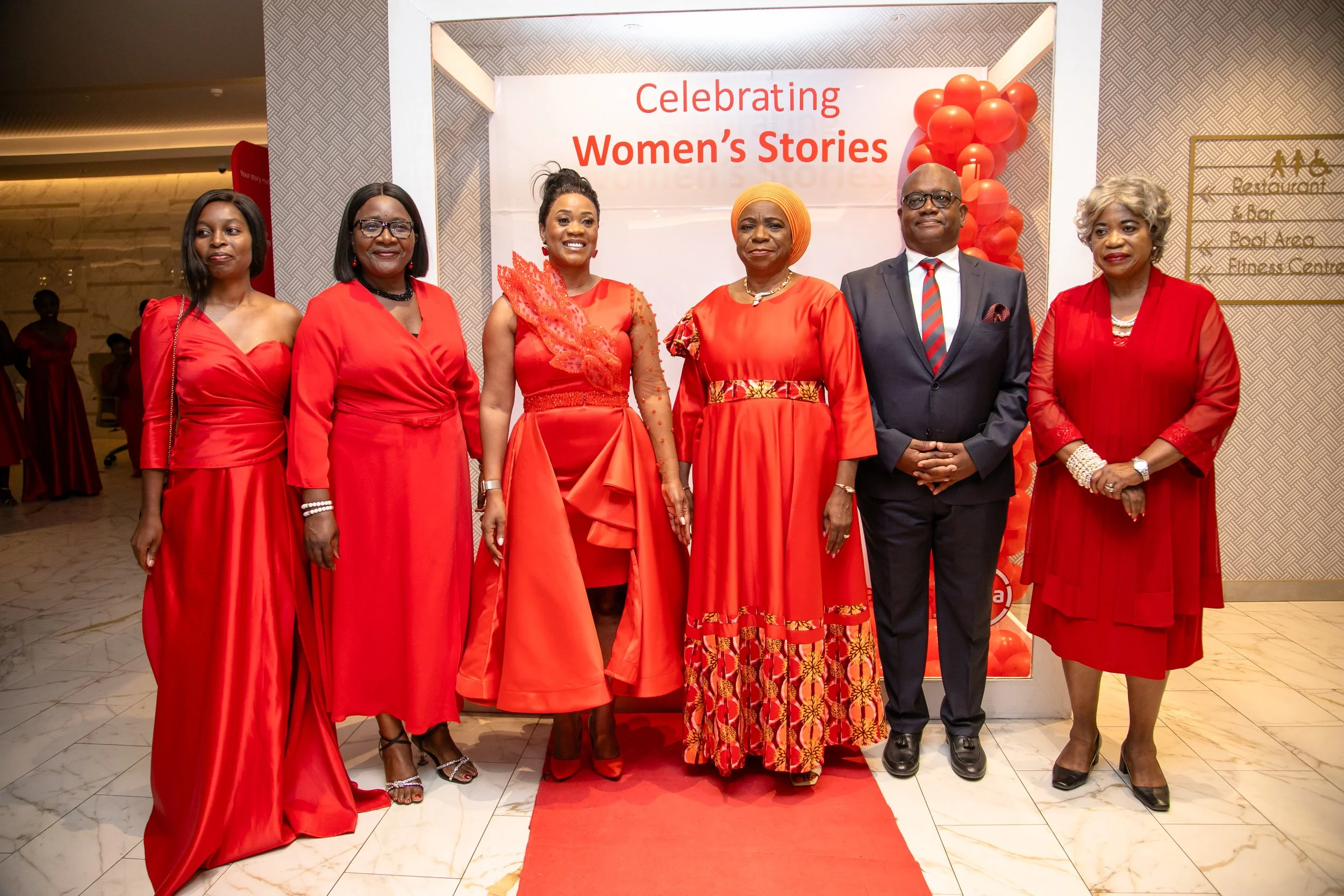 A group of seven women and one man standing in front of a sign that says 'Celebrating Women's Stories'. The women are dressed in elegant red dresses and outfits, and the man is wearing a suit with a striped tie. They are posing for a photo at an even
