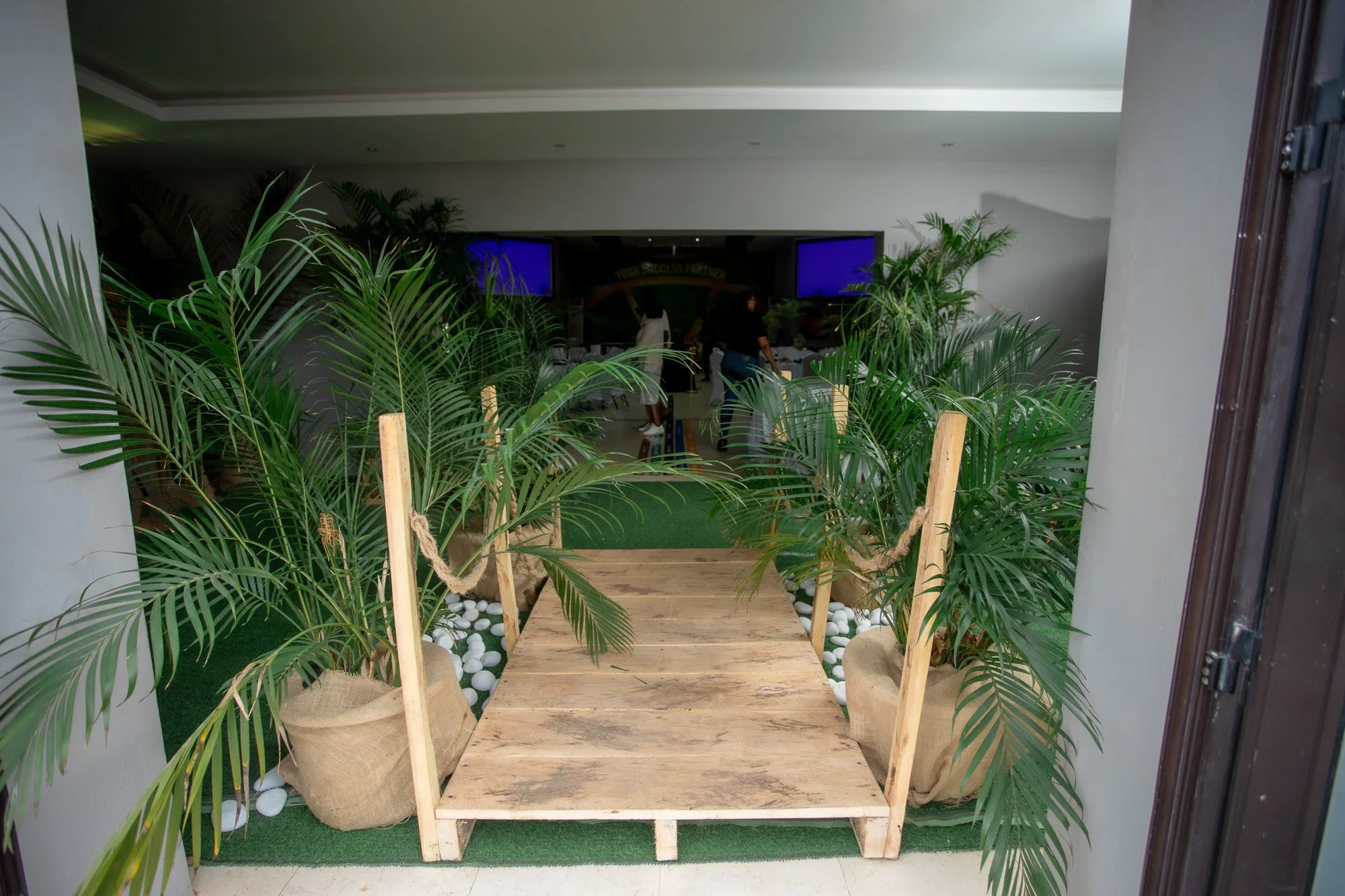 Wooden pathway leading into a room decorated with lush green plants on either side, with white pebbles at the base of the plants and a green carpet underneath.