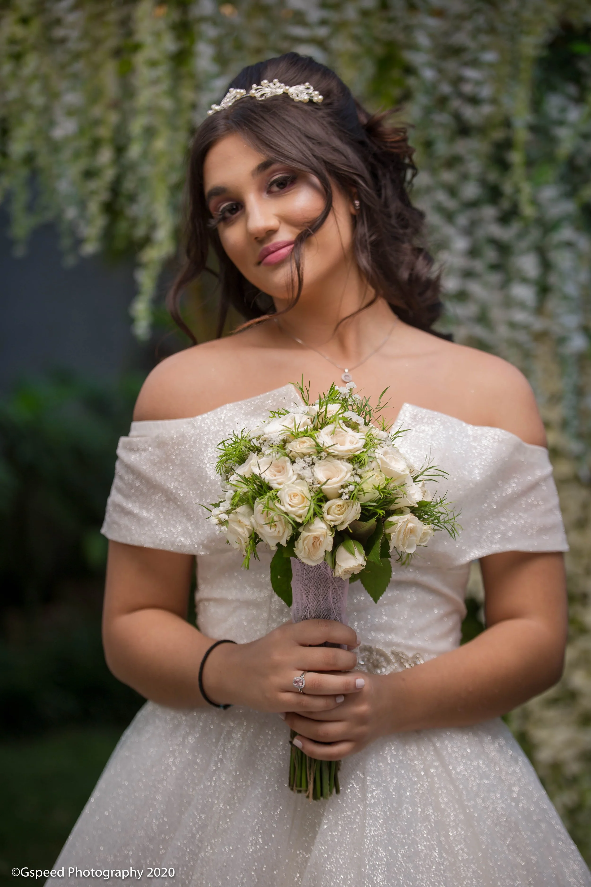 A bride in an off-shoulder, sparkly wedding dress holding a bouquet of white roses with greenery, standing outdoors in front of a blurred green background.