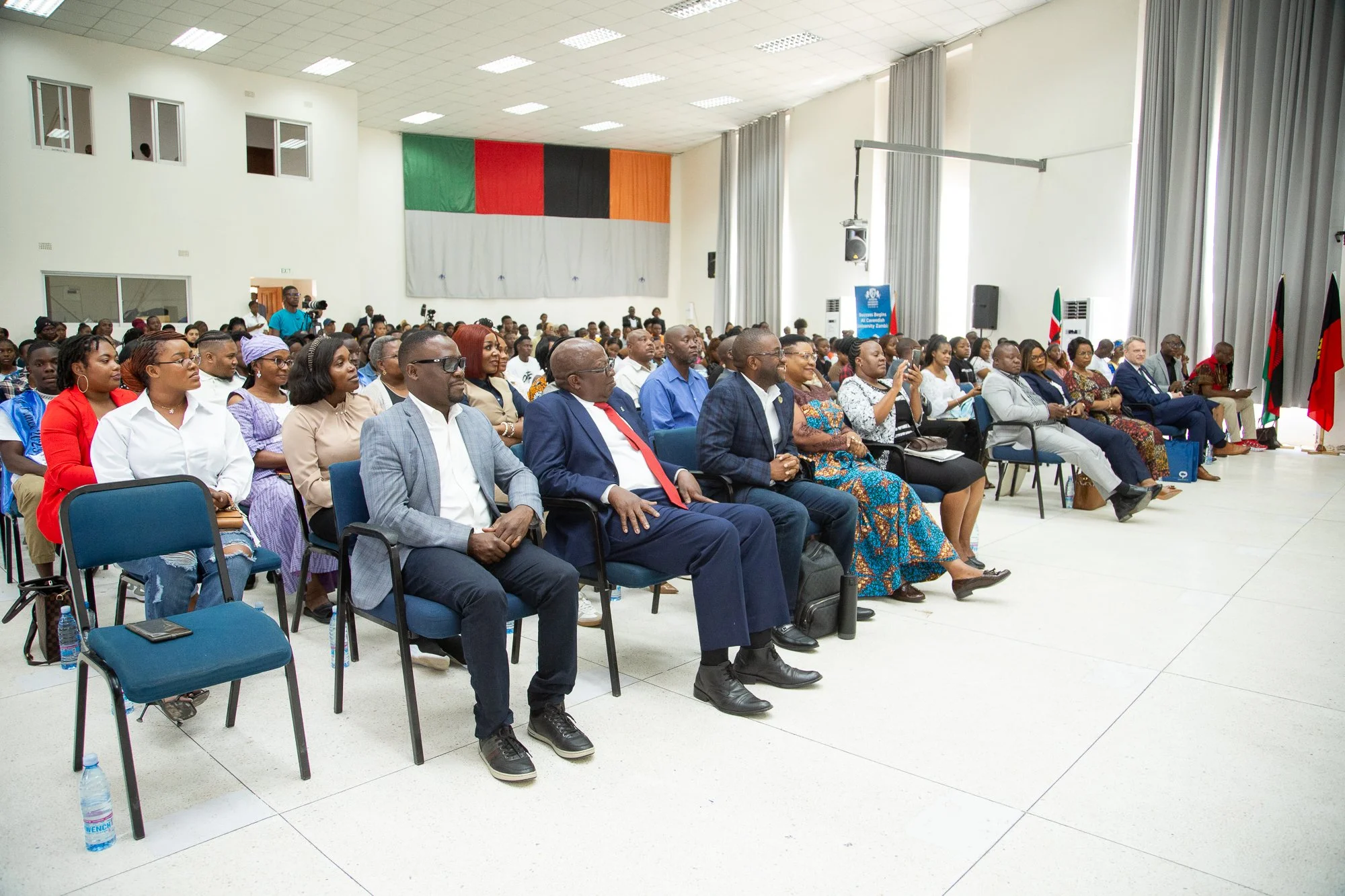 A large group of diverse people seated in a conference hall, attending an event. The room has white walls, high windows, gray curtains, and flags on the right side. The audience includes men and women dressed in formal and traditional attire, with so