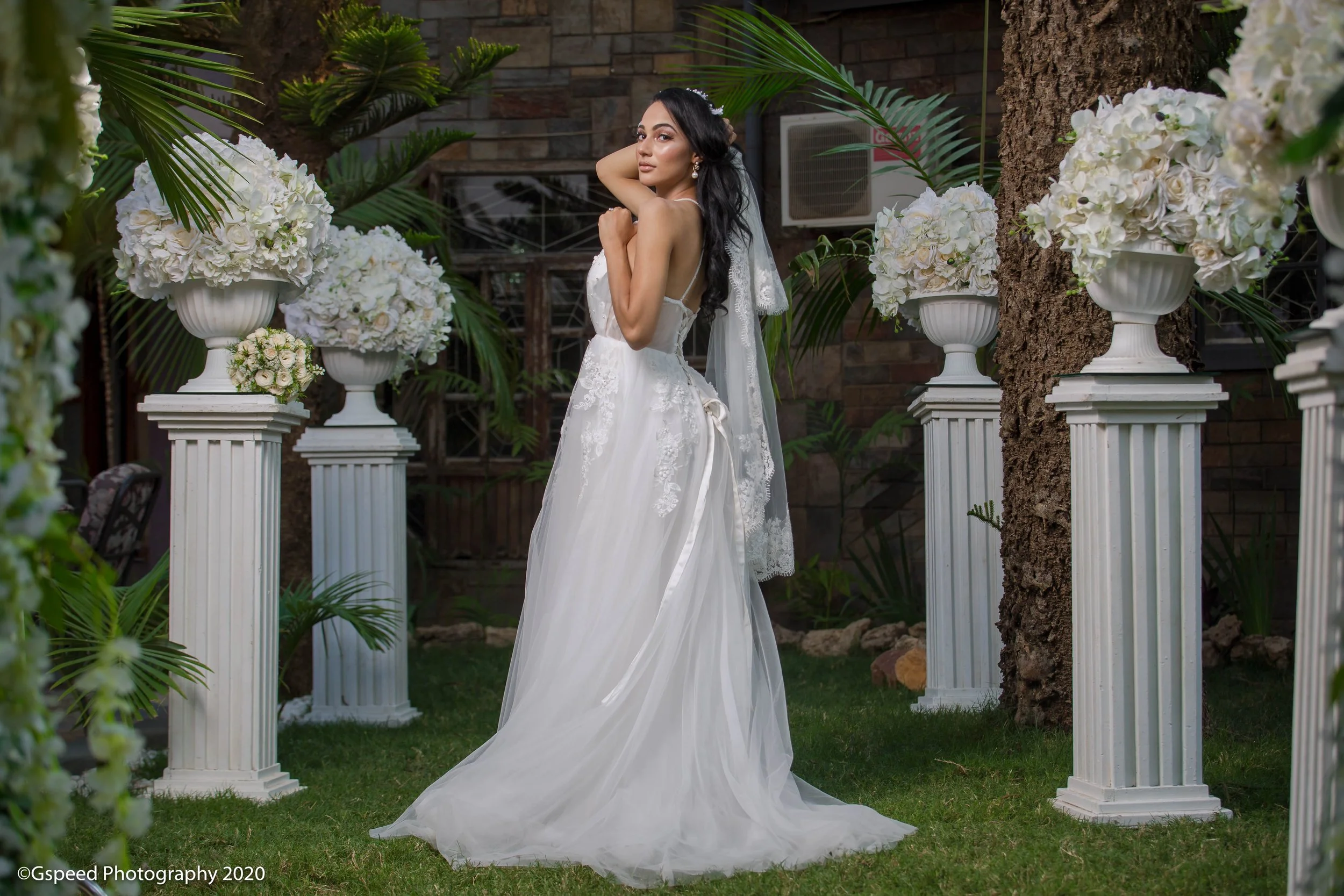 A bride wearing a white wedding dress with lace details, standing outdoors among tall white floral arrangements on pedestals and green foliage, with a brick wall and trees in the background.