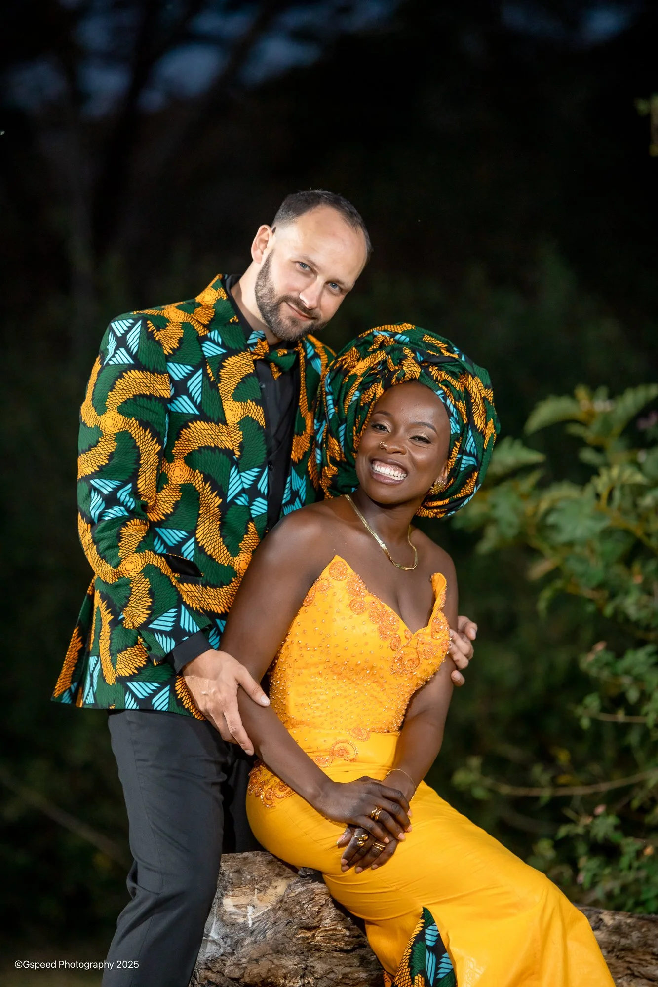 A man with light skin and a beard, and a woman with dark skin and a colorful headwrap, smiling together outdoors at night. The woman is sitting on a log, wearing a bright orange dress, and the man is standing behind her, wearing a vibrant patterned j
