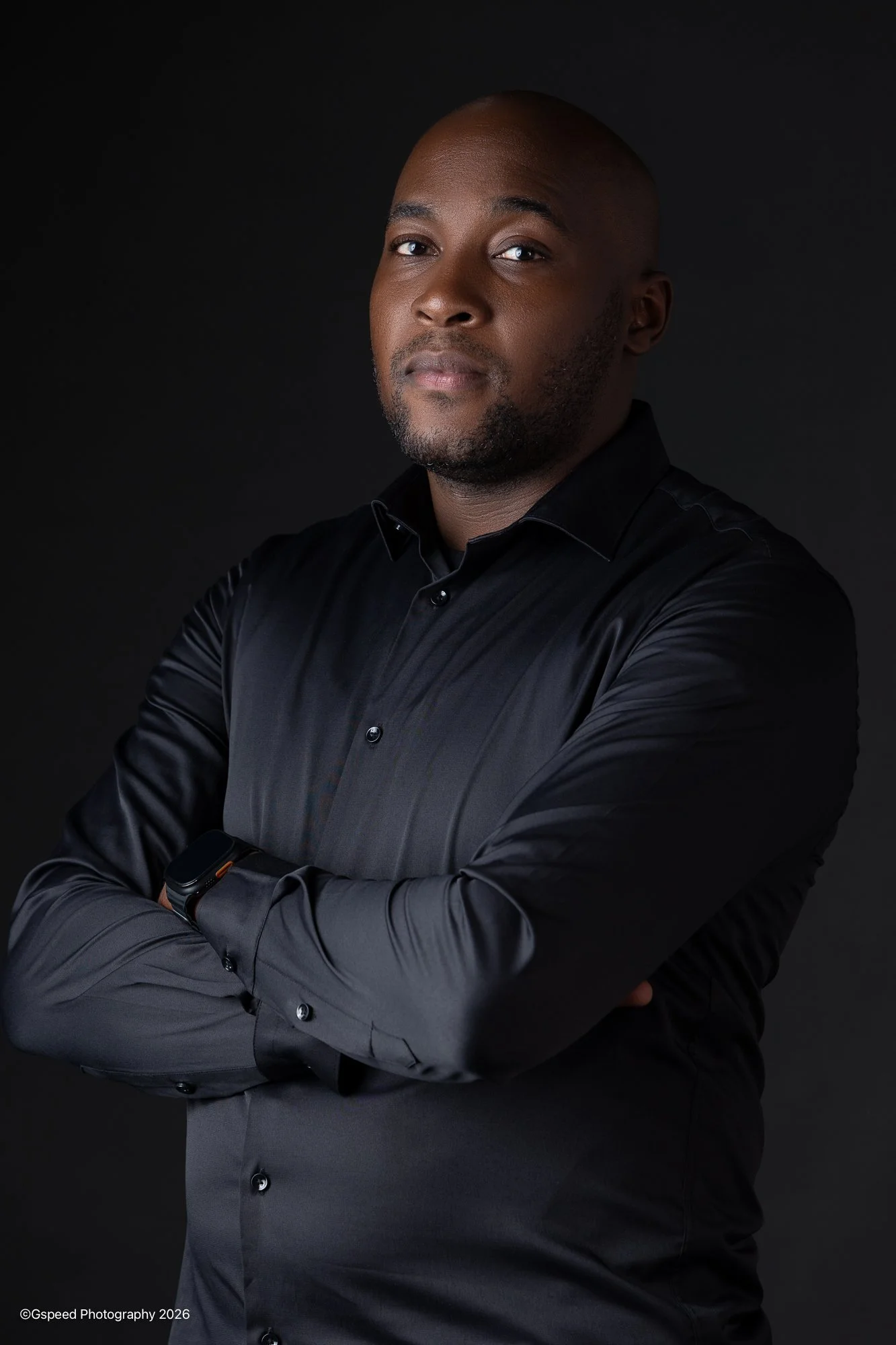 Portrait of a confident man with crossed arms wearing a dark dress shirt and smartwatch against a black background.