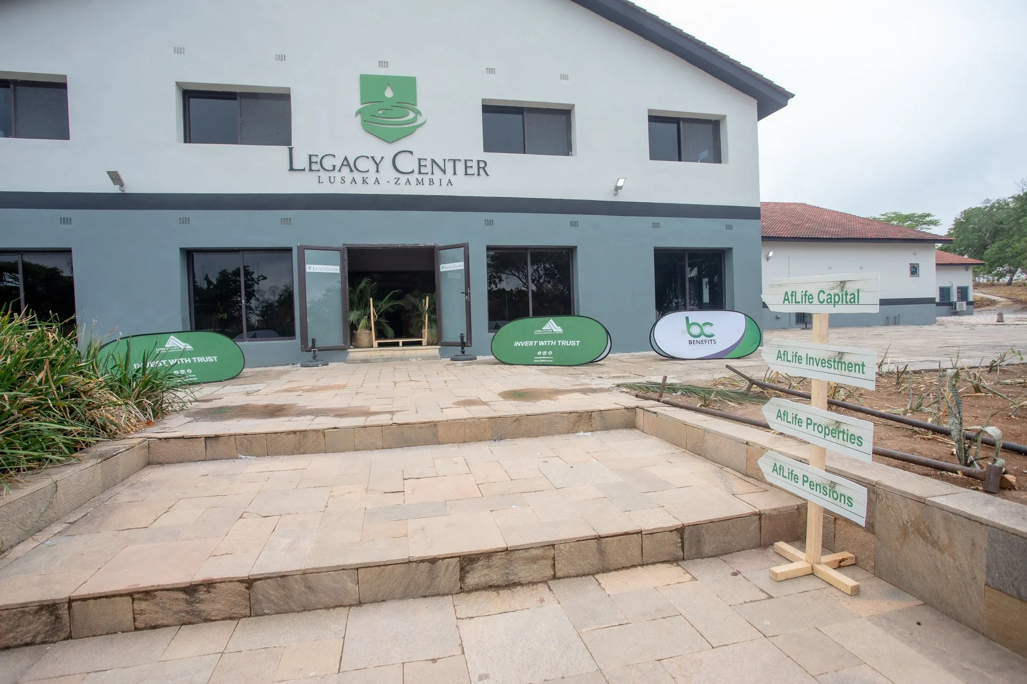 Exterior of a building labeled 'Legacy Center Lusaka-Zambia' with signs for financial services including investments, benefits, properties, and pensions. The building has a white facade with multiple windows and a logo featuring a water droplet and a