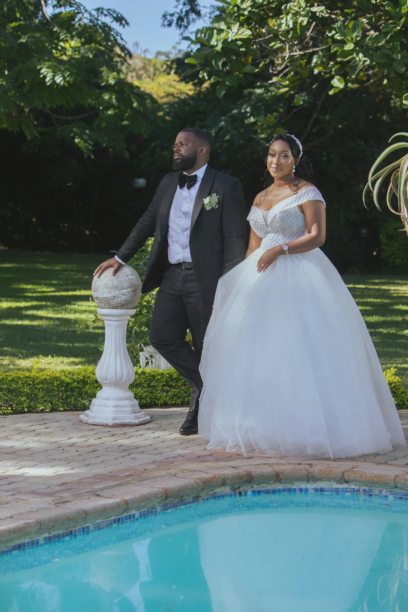 A bride and groom dressed in wedding attire, standing outdoors by a pool with green trees behind them. The groom leans on a white pedestal with a globe, and the bride stands beside him in a white wedding gown, both posing for a wedding photo.