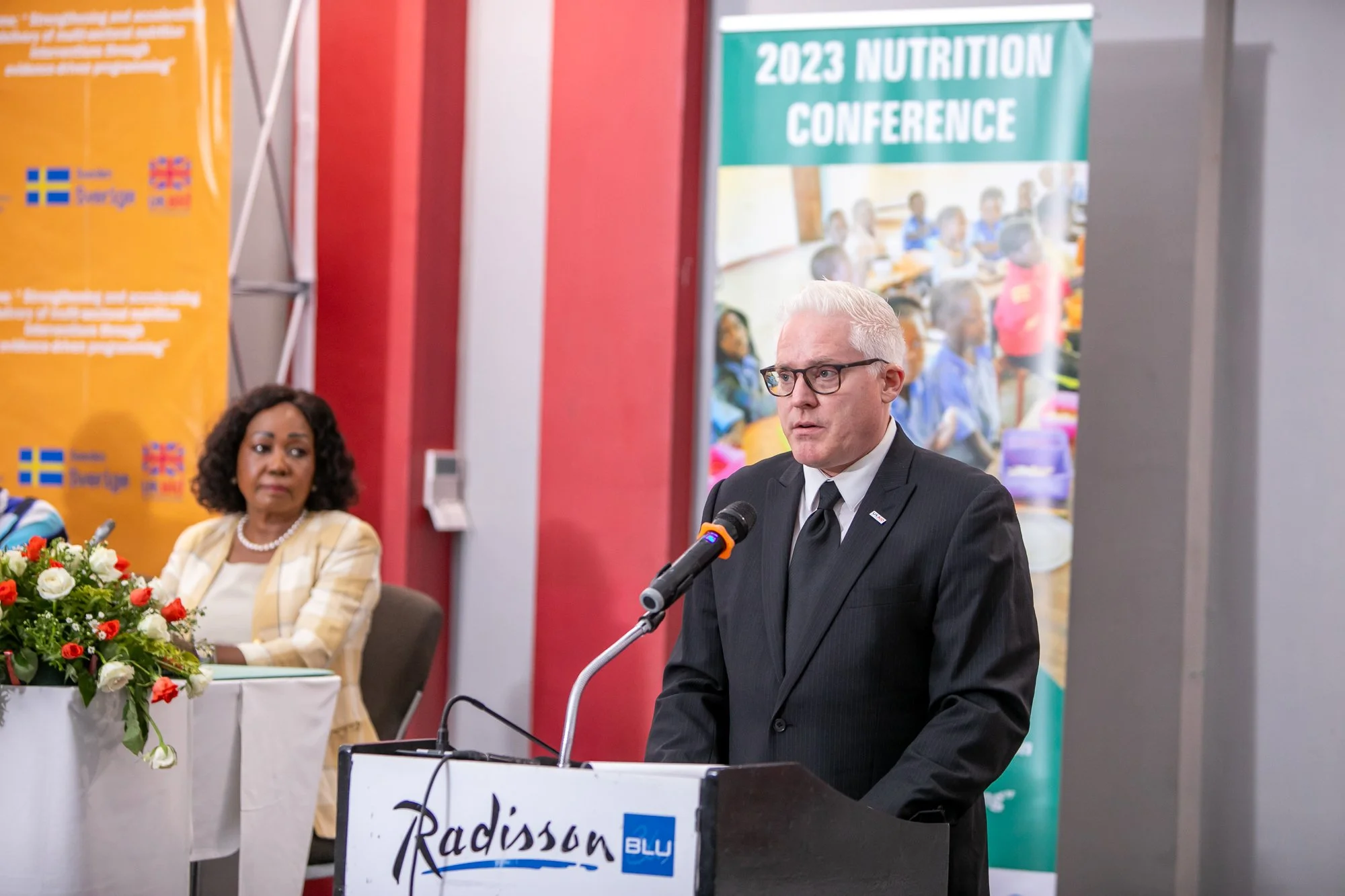 A man in a dark suit and glasses speaking at a microphone during a conference, with a woman in a yellow striped blazer sitting beside him. There are banners in the background reading '2023 Nutrition Conference' and 'Radisson Blu'. A bouquet of flower