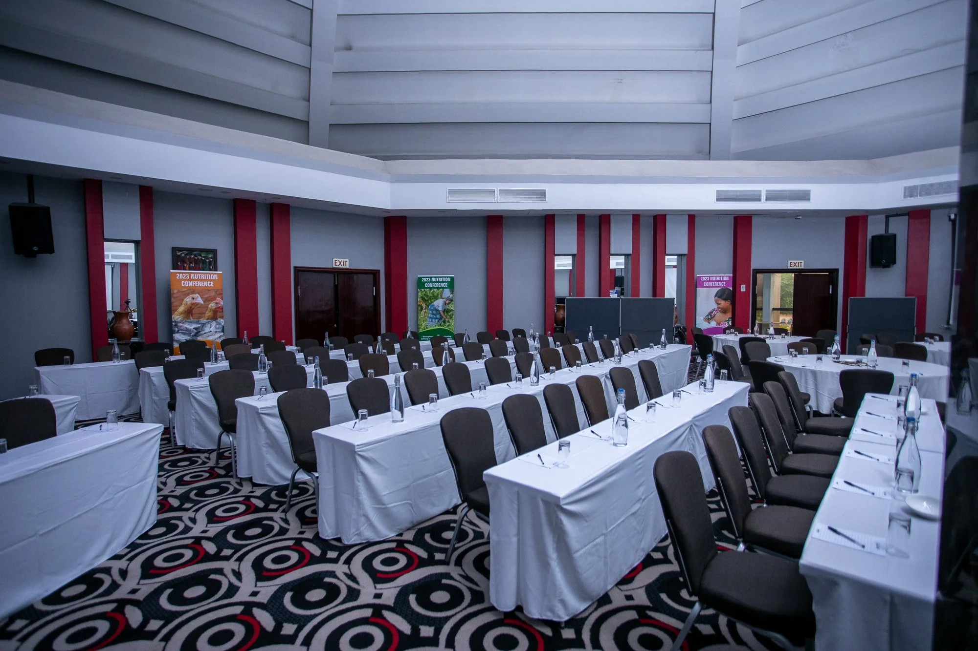 Conference room set up with long tables covered in white tablecloths, paired with chairs, and water bottles placed on each table. The room has patterned carpet flooring, red accents on the walls, and banners that read '2023 Nutrition Conference.' The