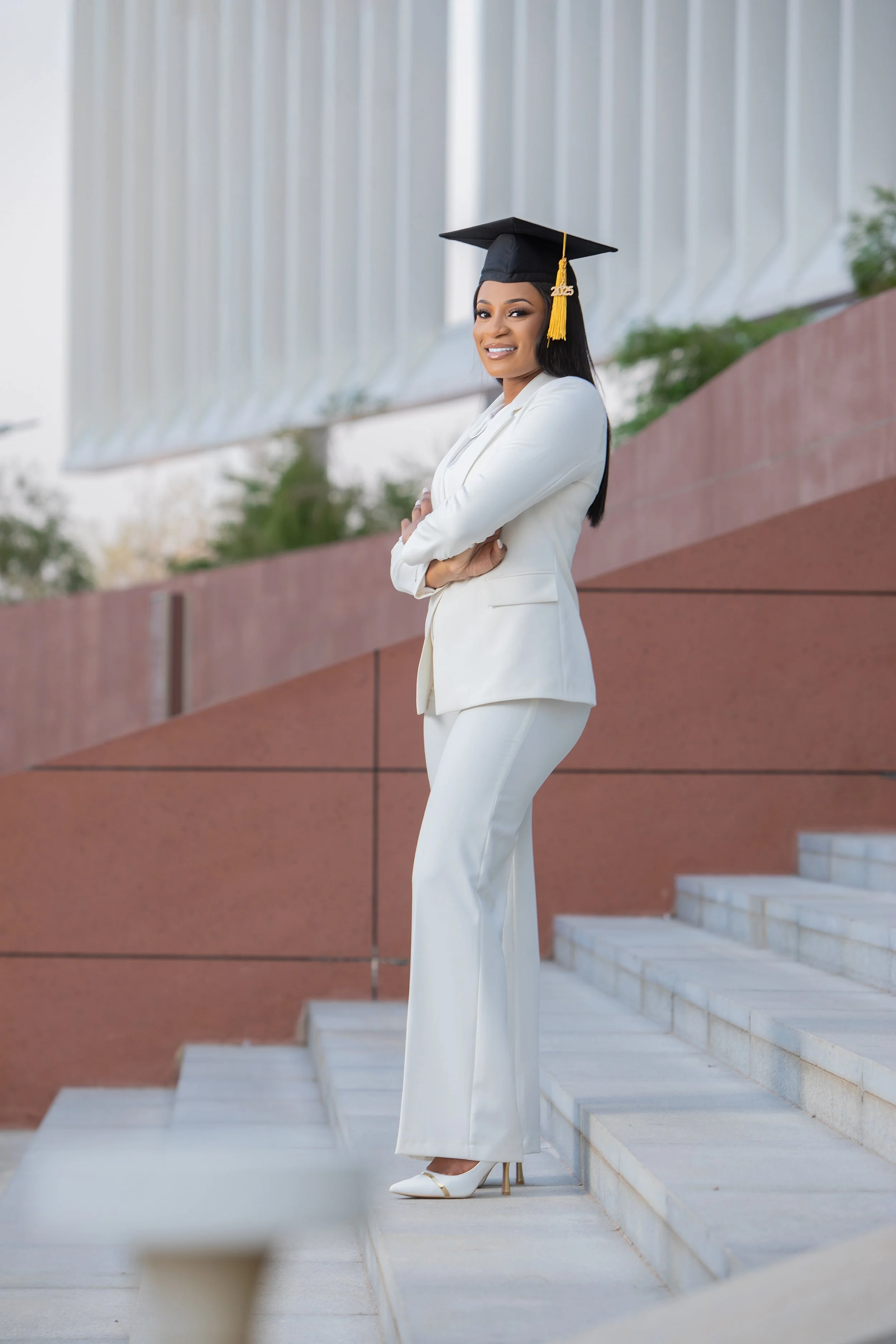 A young woman in a white pantsuit and white heels, wearing a black graduation cap with a yellow tassel that reads '2025,' standing on outdoor stairs, smiling with her arms crossed.