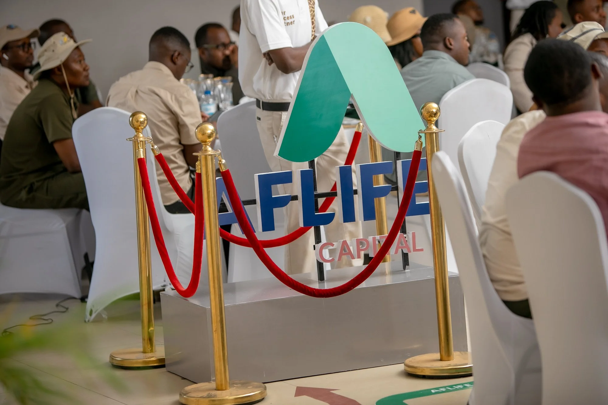 People sitting at a conference or seminar with a large green and blue logo sign in the foreground. The sign reads 'CAPITAL' and is surrounded by red velvet ropes. The attendees appear to be listening attentively, with some wearing hats and glasses.