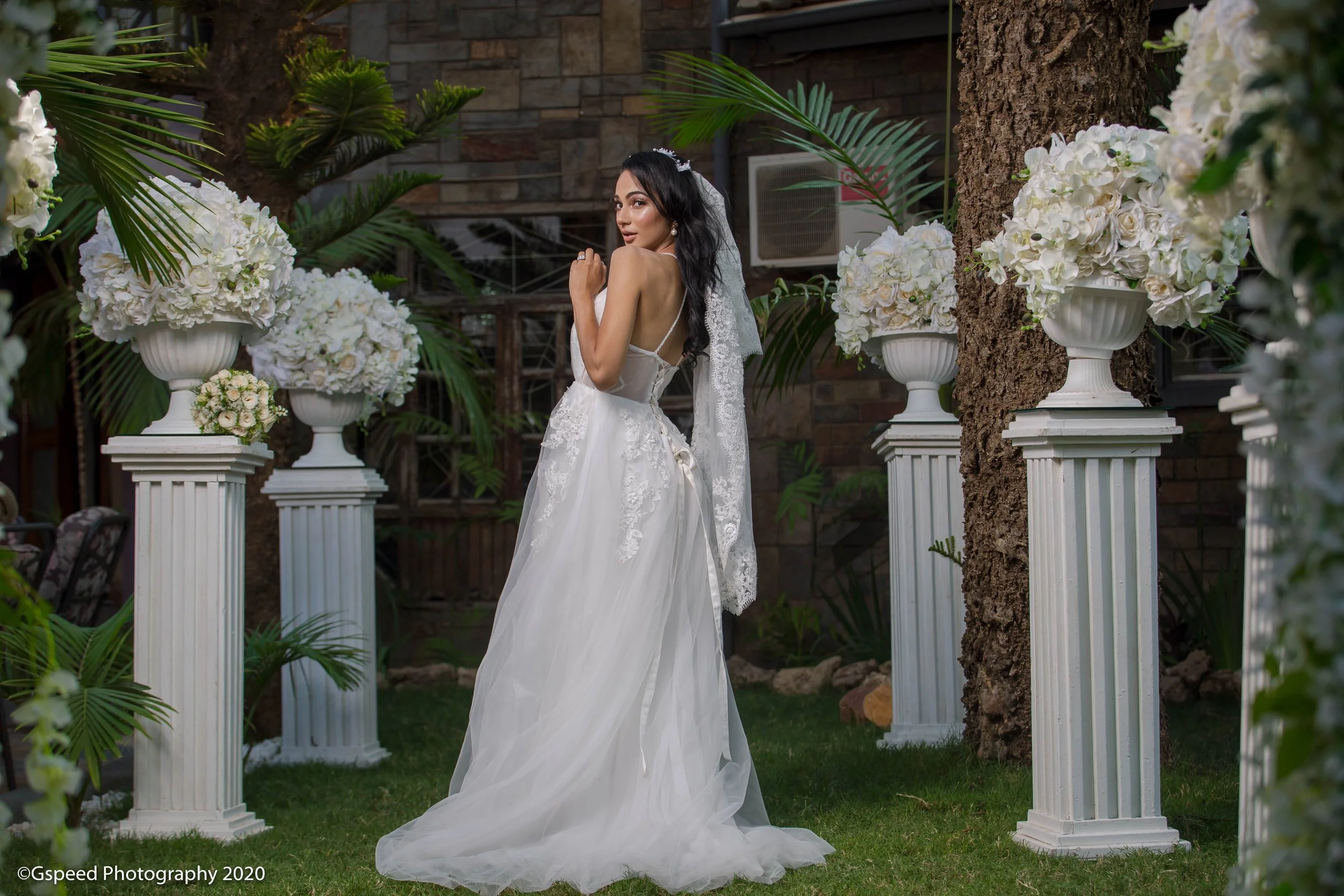 A woman in a white wedding dress with lace details, standing outdoors among white floral arrangements on tall white pedestals, greenery, and trees, looking over her shoulder.