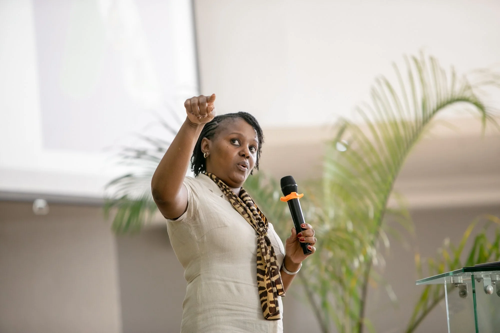 A woman with short curly hair holding a microphone and speaking, raising her right hand in a fist gesture, standing indoors with greenery in the background.
