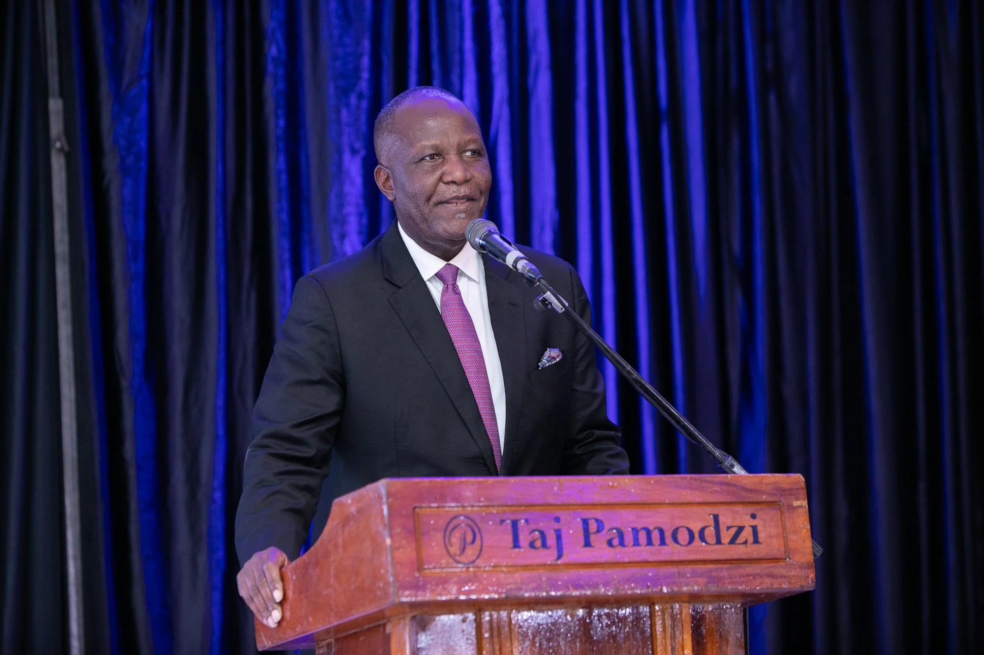 A man in a dark suit and pink tie standing behind a wooden podium with a microphone, speaking at an event with a dark blue curtain backdrop.