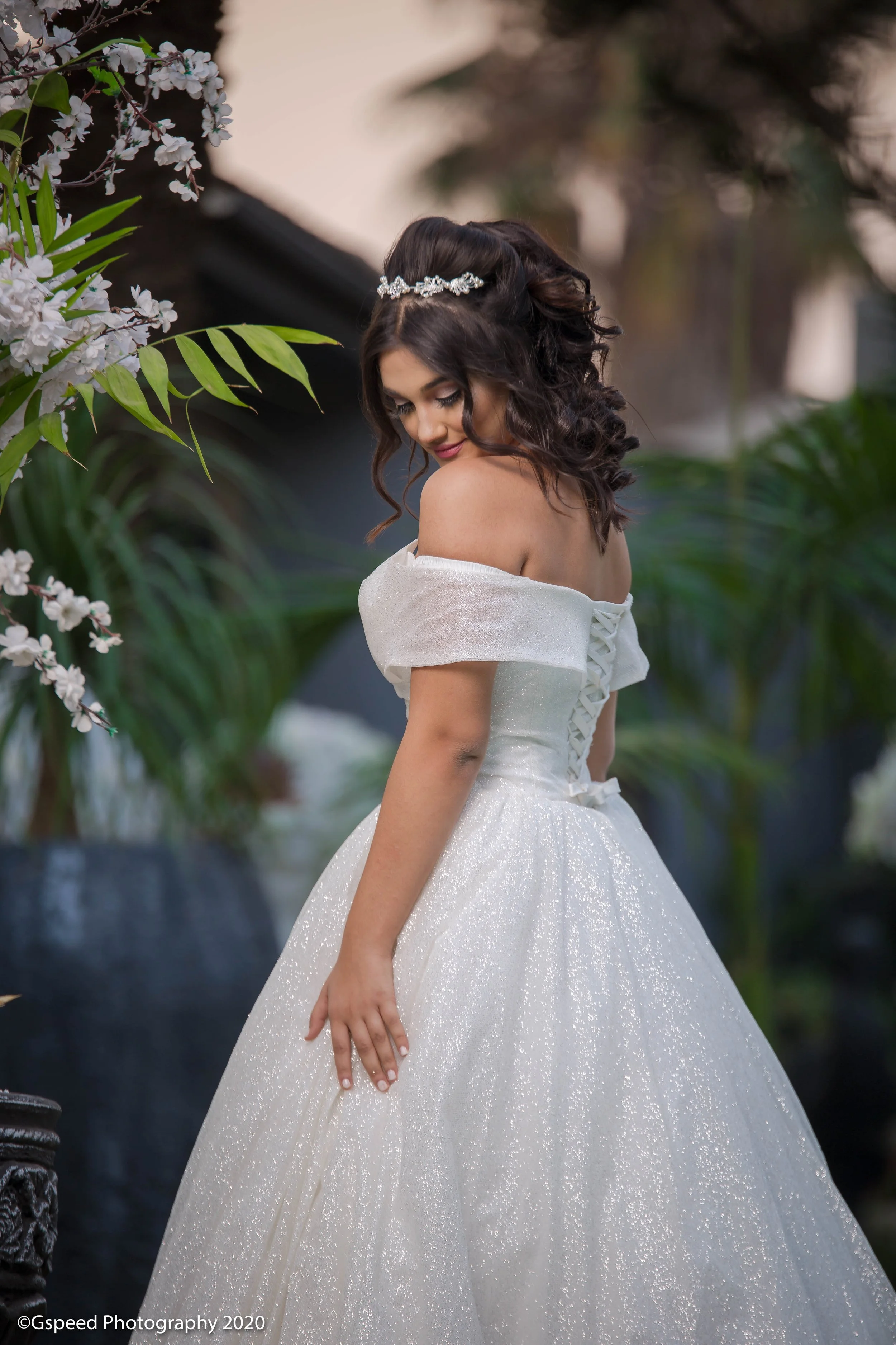 A woman in a white wedding dress with off-the-shoulder sleeves and a lace-up back, standing outdoors surrounded by greenery and white flowers, with a tiara in her dark, curly hair, looking down with a soft smile.