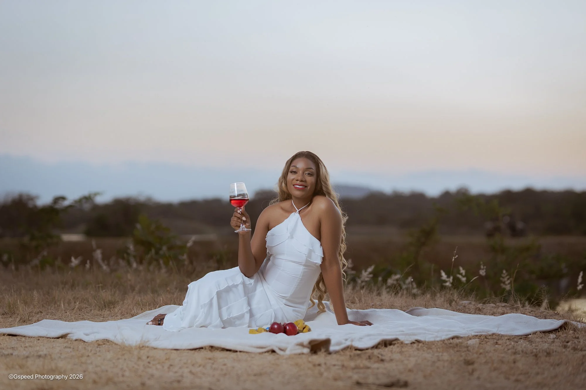 A woman in a white dress sitting on a white blanket outdoors during sunset, holding a glass of red wine, with a small spread of fruit including apples and bananas in front of her, with trees and an open landscape in the background.