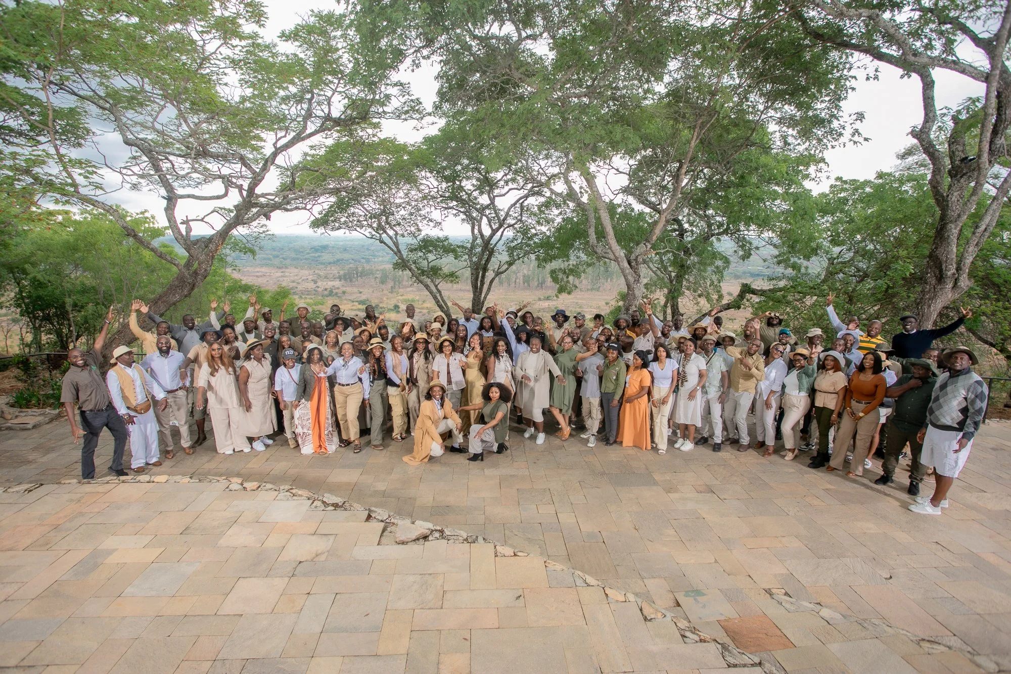 A large group of people standing outdoors on a paved area under trees, posing for a group photo. The background shows a scenic landscape with trees and hills.