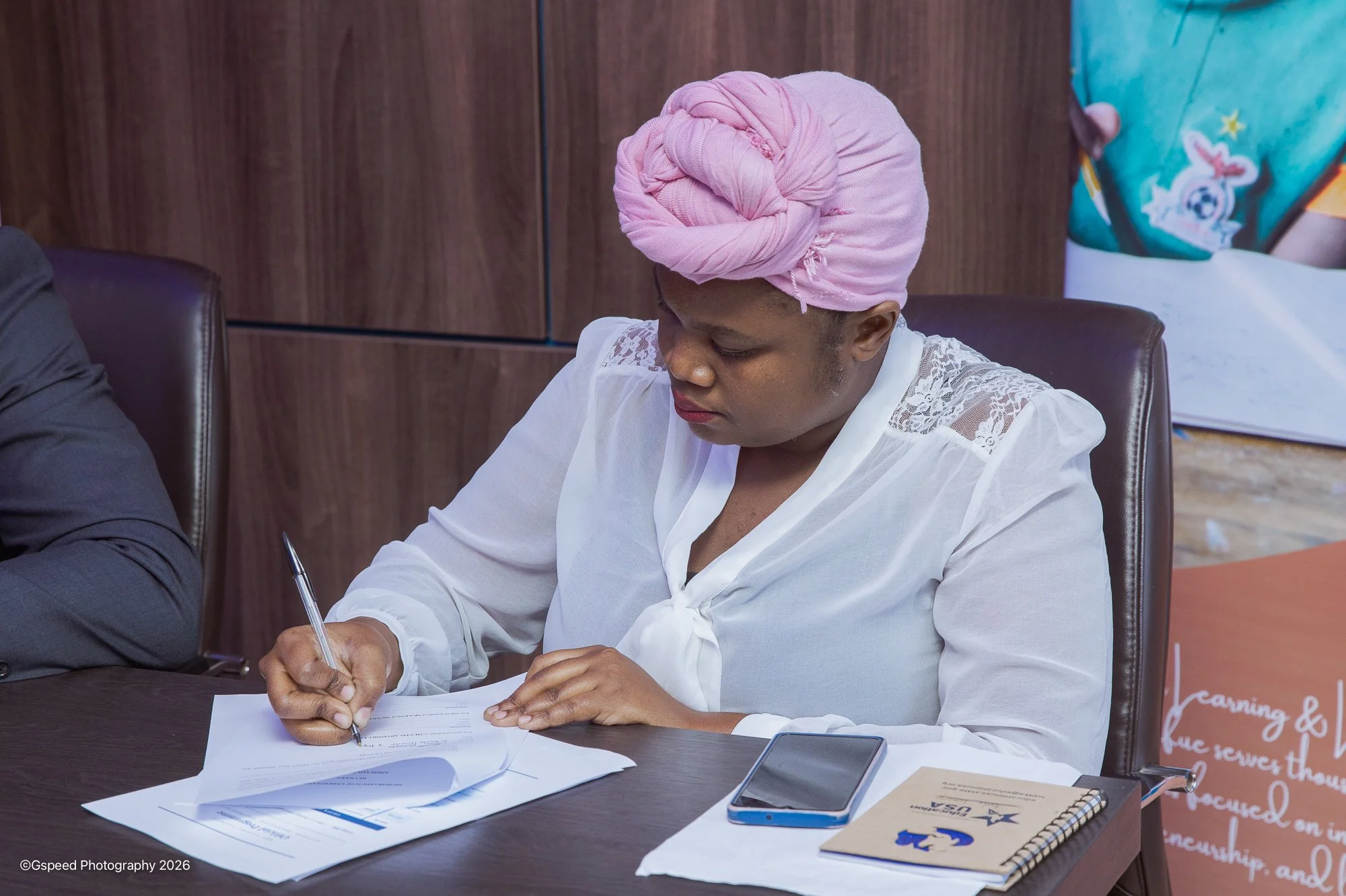 A woman with a pink headwrap, white lace top, and makeup sitting at a desk, writing on a document with a pen. On the desk, there are papers, a smartphone, and a notebook. A person next to her is partially visible, and behind her, there is a wooden wa