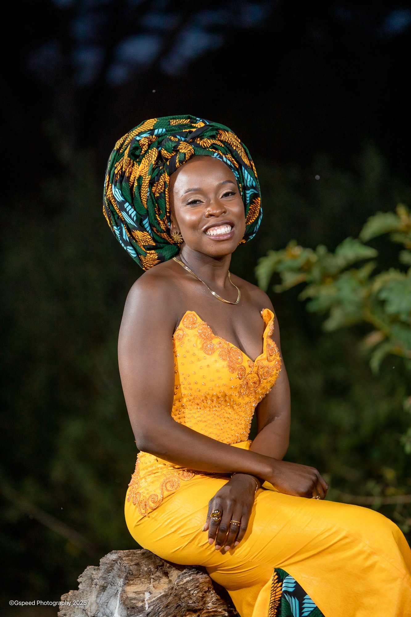 A woman smiling and sitting on a tree stump outdoors at night, wearing a bright yellow dress with orange embroidery and a colorful headwrap with green, yellow, black, and blue patterns.
