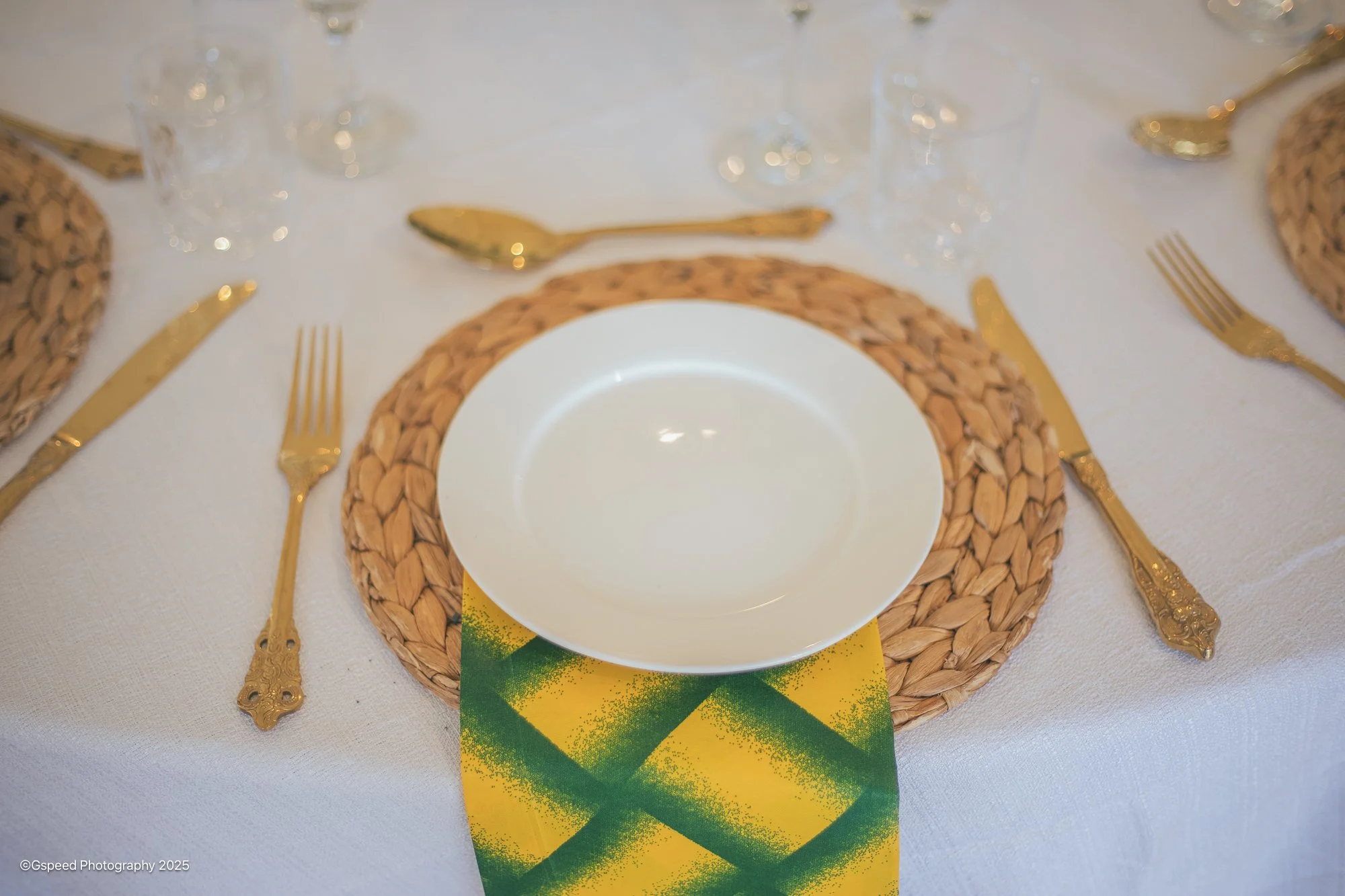 A table setting with a white plate on a round woven placemat, gold flatware including a fork, knife, and spoon, wine glasses, and a colorful napkin with green and yellow colors underneath the plate.