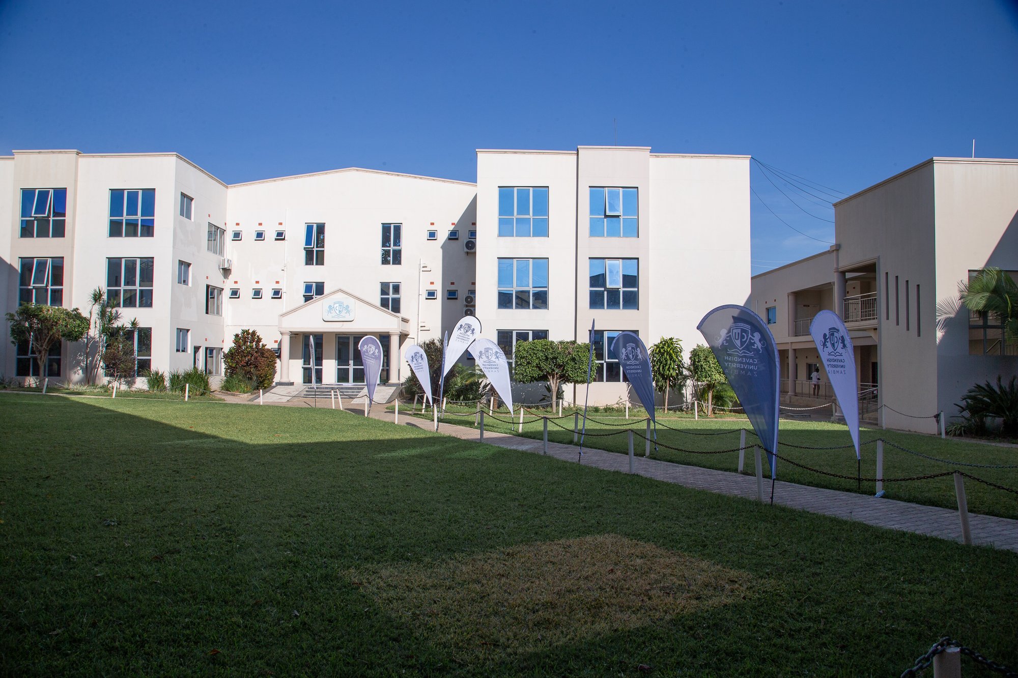 White multi-story building with blue windows and a small entrance, surrounded by green grass and trees, with university flags in front.