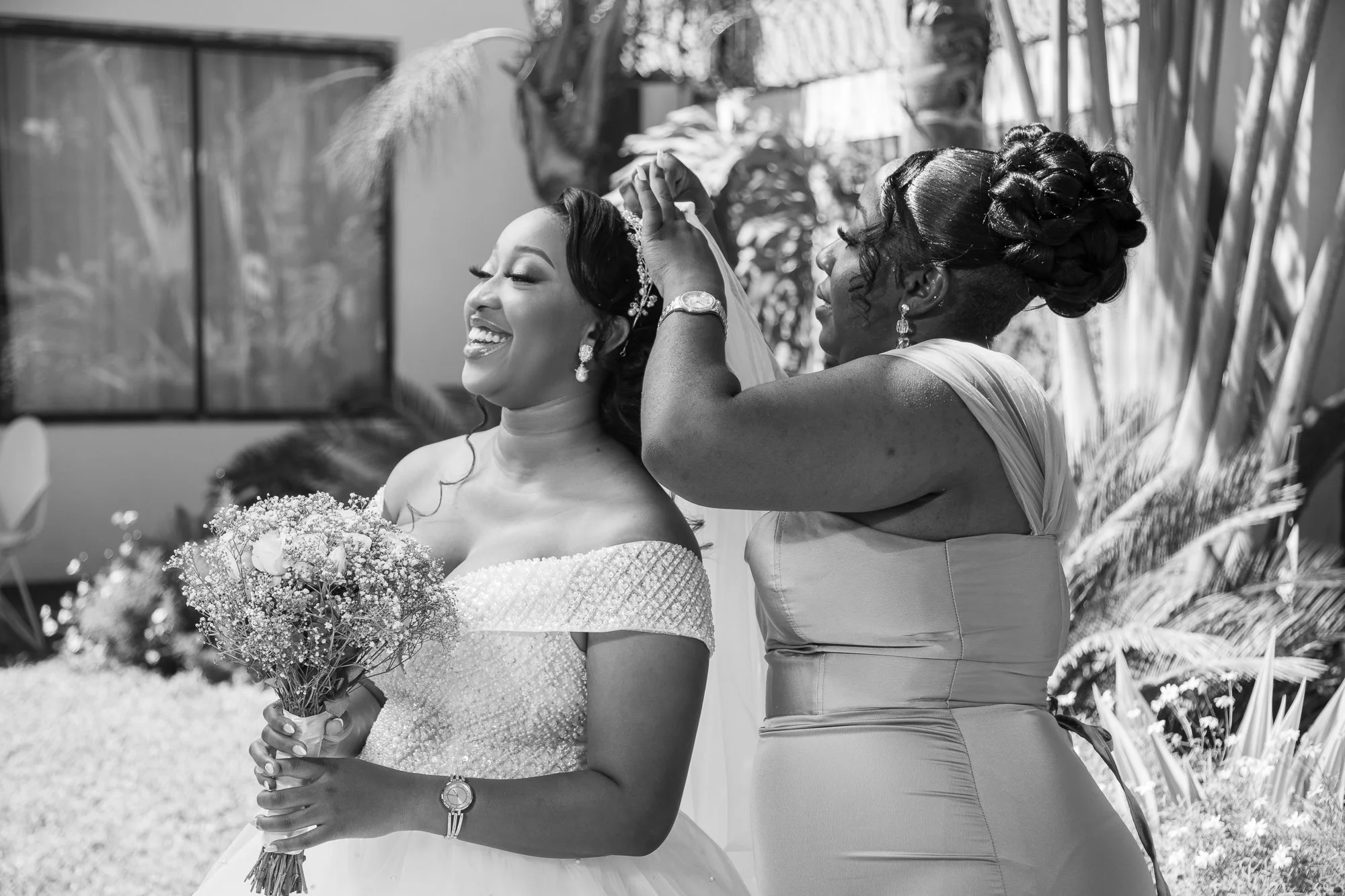 A woman in wedding attire smiling as another woman places a tiara on her head during a wedding ceremony. The woman in wedding attire holds a bouquet of flowers and wears a dress with lace details. The scene is outdoors with trees and plants in the ba