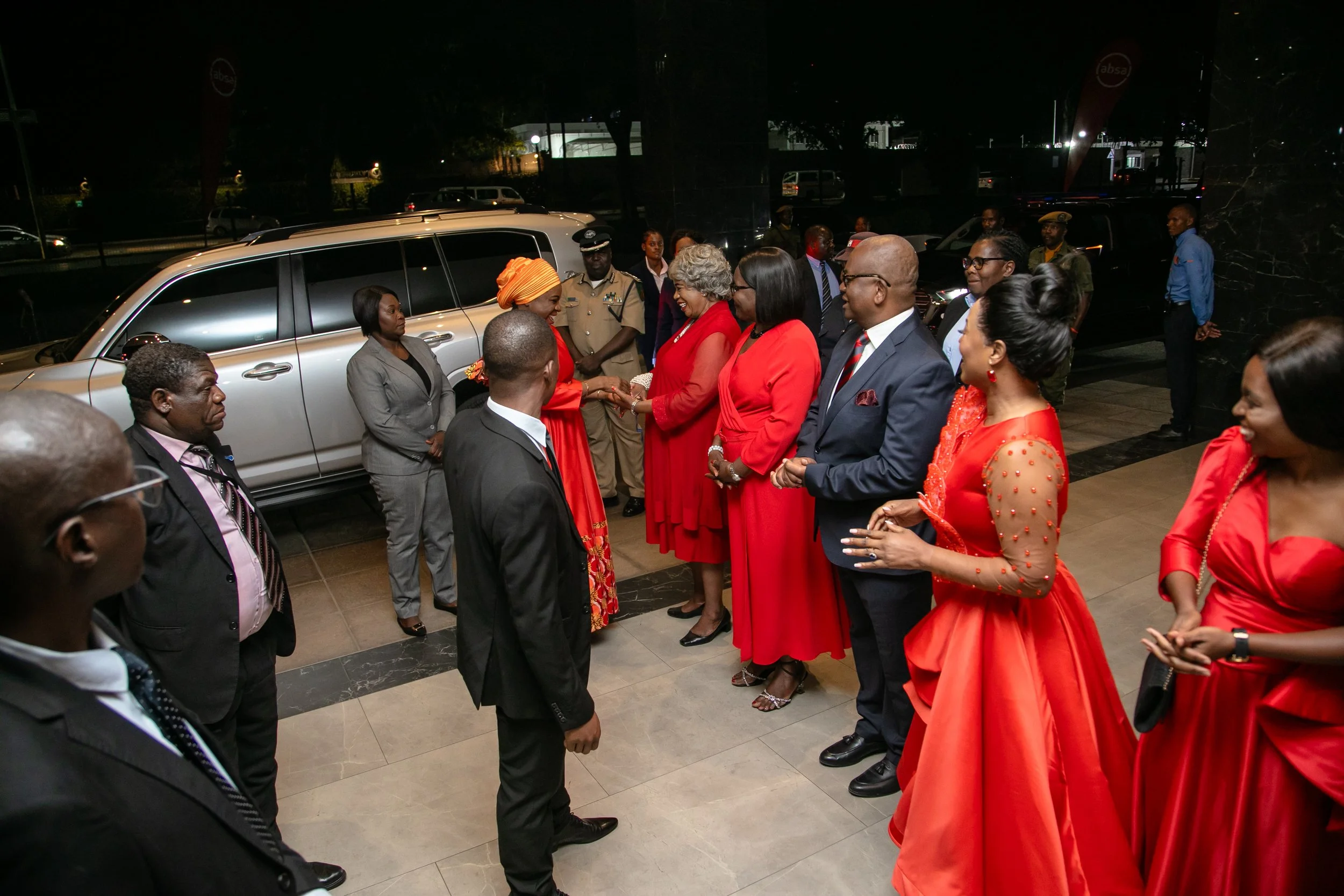 A group of people, including women in red dresses and men in suits, greet another woman in a red dress at an indoor event. A woman in a grey suit and a woman in an orange headwrap are also part of the greeting. Police officers and other attendees are