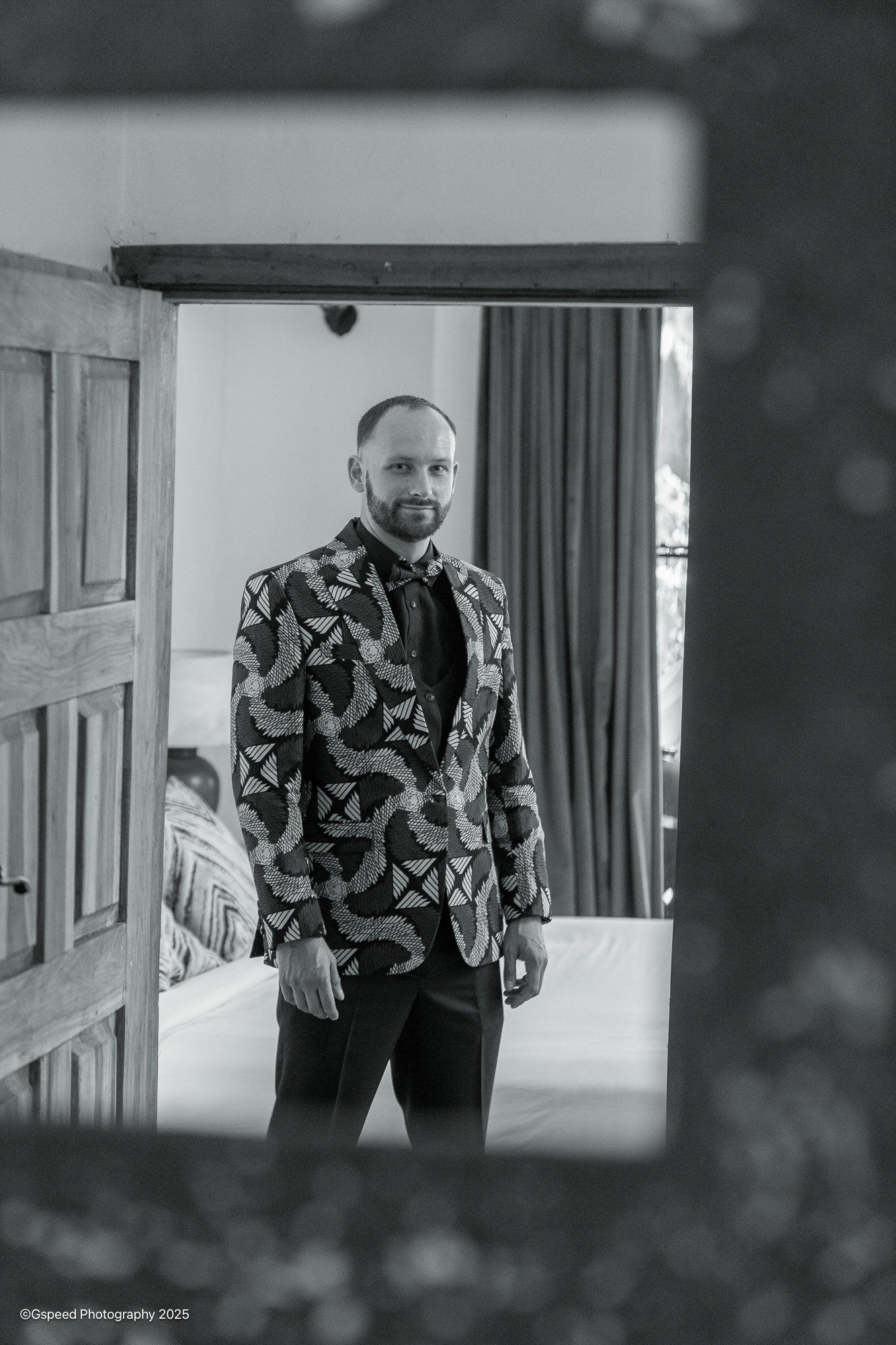 A man with a beard and short hair wearing a patterned suit jacket and bow tie, standing in a room next to a bed with striped bedding, as seen through a decorative mirror or frame.