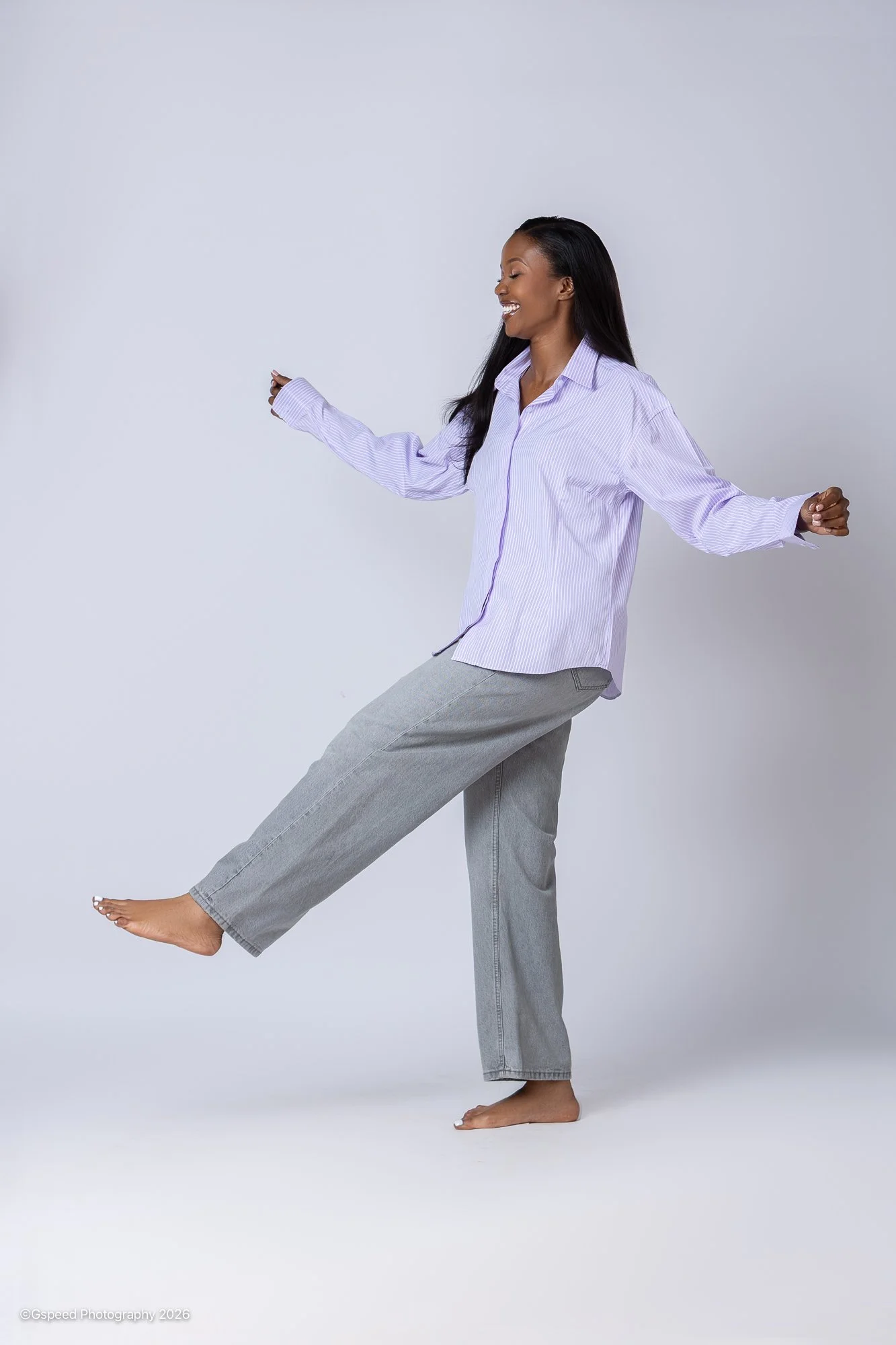A woman with dark hair, wearing a light purple striped button-up shirt and gray pants, balancing on one leg with eyes closed and a joyful expression against a plain light gray background.