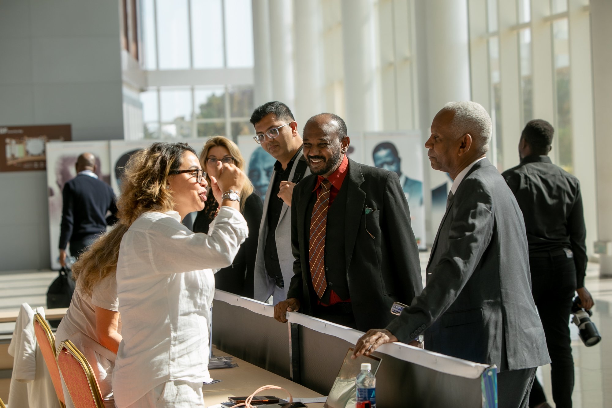 A diverse group of professionals engaged in a friendly conversation at a conference or event in a modern, well-lit building with large windows. A woman seated at a table is talking animatedly while three men stand nearby listening and smiling.