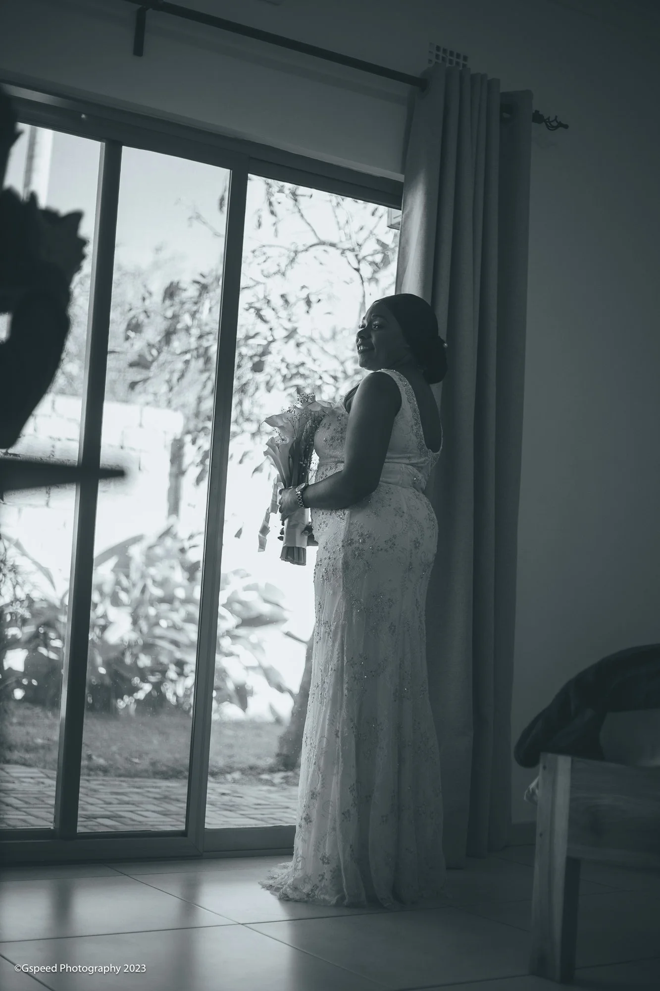 A woman in a wedding dress holding a bouquet standing inside by a glass door with snow outside.