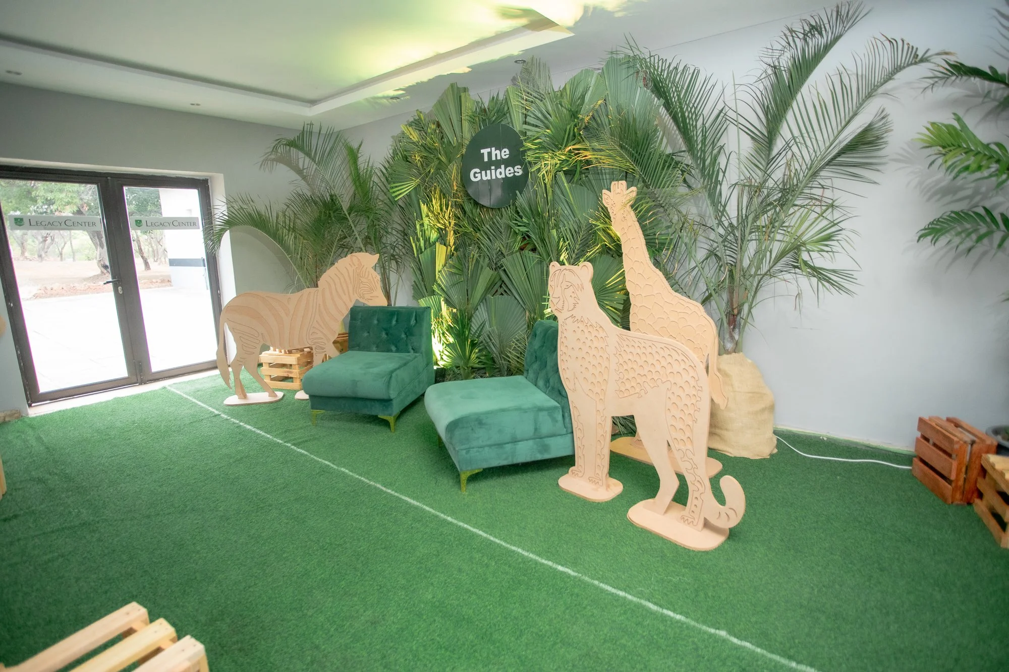 Indoor space with a green carpeted floor, tropical plants on the wall, and cardboard cutouts of jungle animals including a giraffe, lion, and tiger. There are two green chairs and a window door with the sign 'Legacy Center' on it.