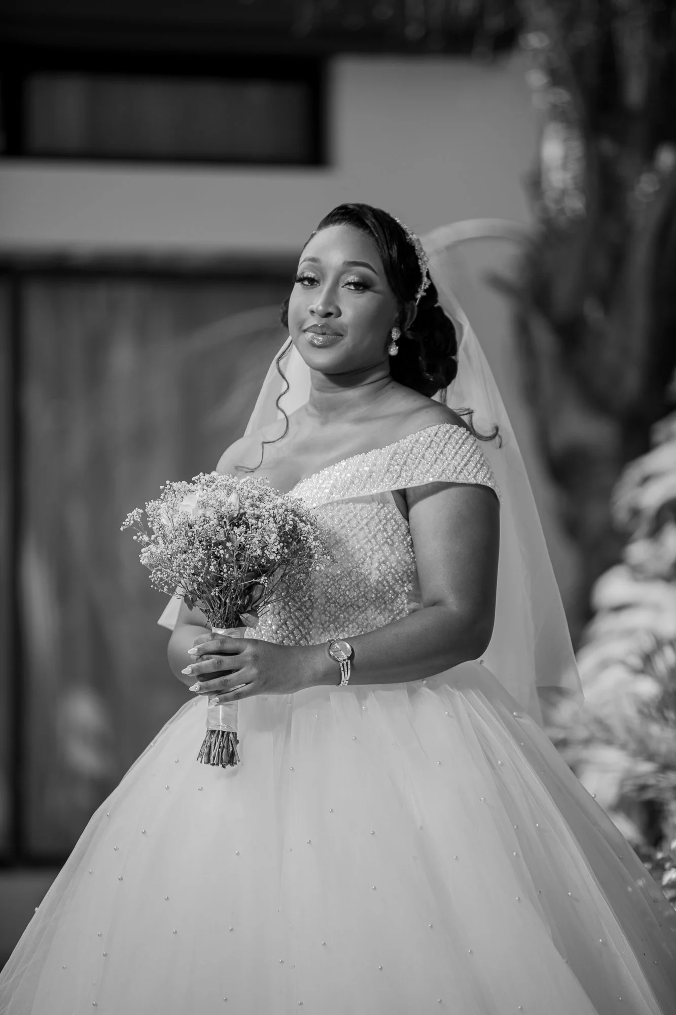 A Black and White Photo of a Bride with a Wedding Dress, Veil, Earrings, and a Watch, holding a bouquet of flowers.