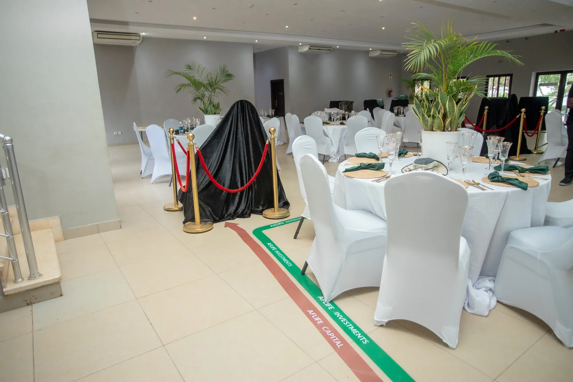 A banquet hall with round tables covered in white tablecloths and set with glassware and napkins. Large potted plants decorate the room. The floor has a marked line labeled 'Alife Investments' with red and green colors, possibly indicating social dis