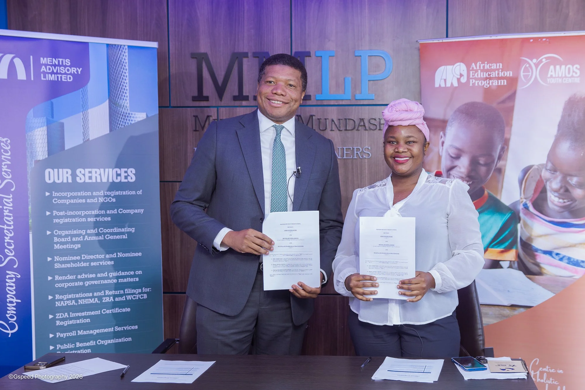 A man in a dark suit and a woman in a white blouse with a pink headwrap are smiling and holding documents in a professional setting. Behind them are banners and a large sign with the letters 'M.I.L.P' indicating an organization or event related to Mu