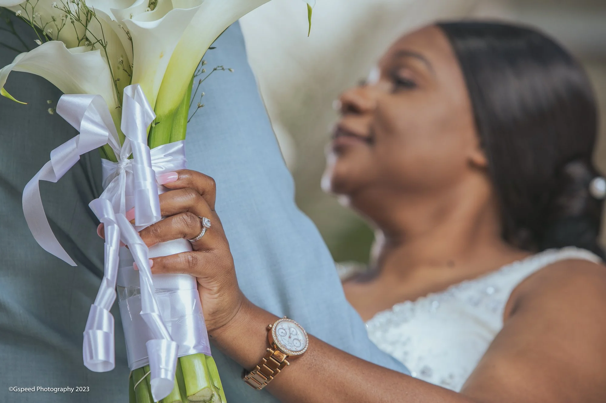 A woman in a white dress holds a bouquet of white calla lilies with ribbon, and wears a gold wristwatch and ring, smiling at an outdoor event.