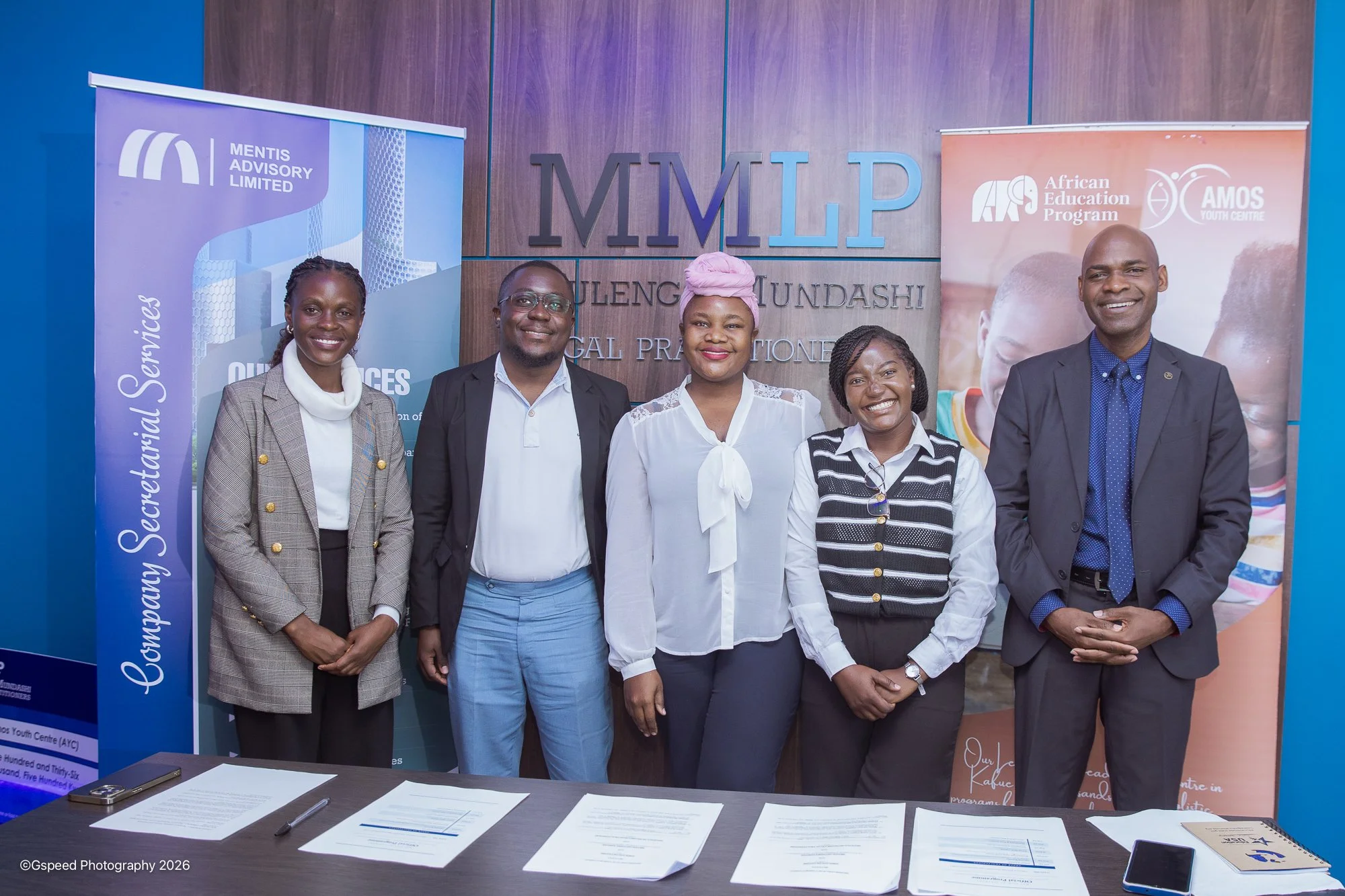 Five people standing in front of a wall with logos and the text 'MMLP'. They are smiling and dressed in business attire. Two banners are visible, one with a blue background on the left and one with a peach background on the right. A table with papers